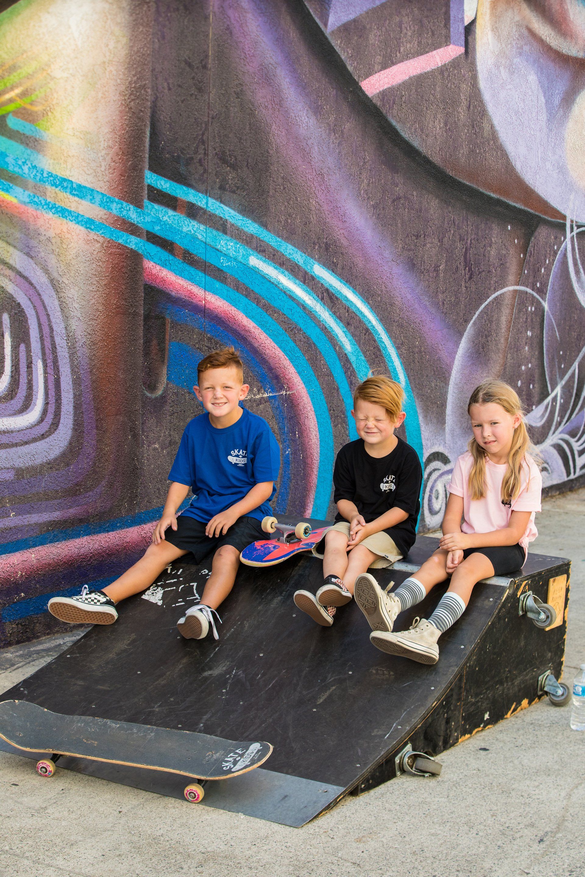 Three children are sitting on skateboards on a ramp in front of a graffiti wall.