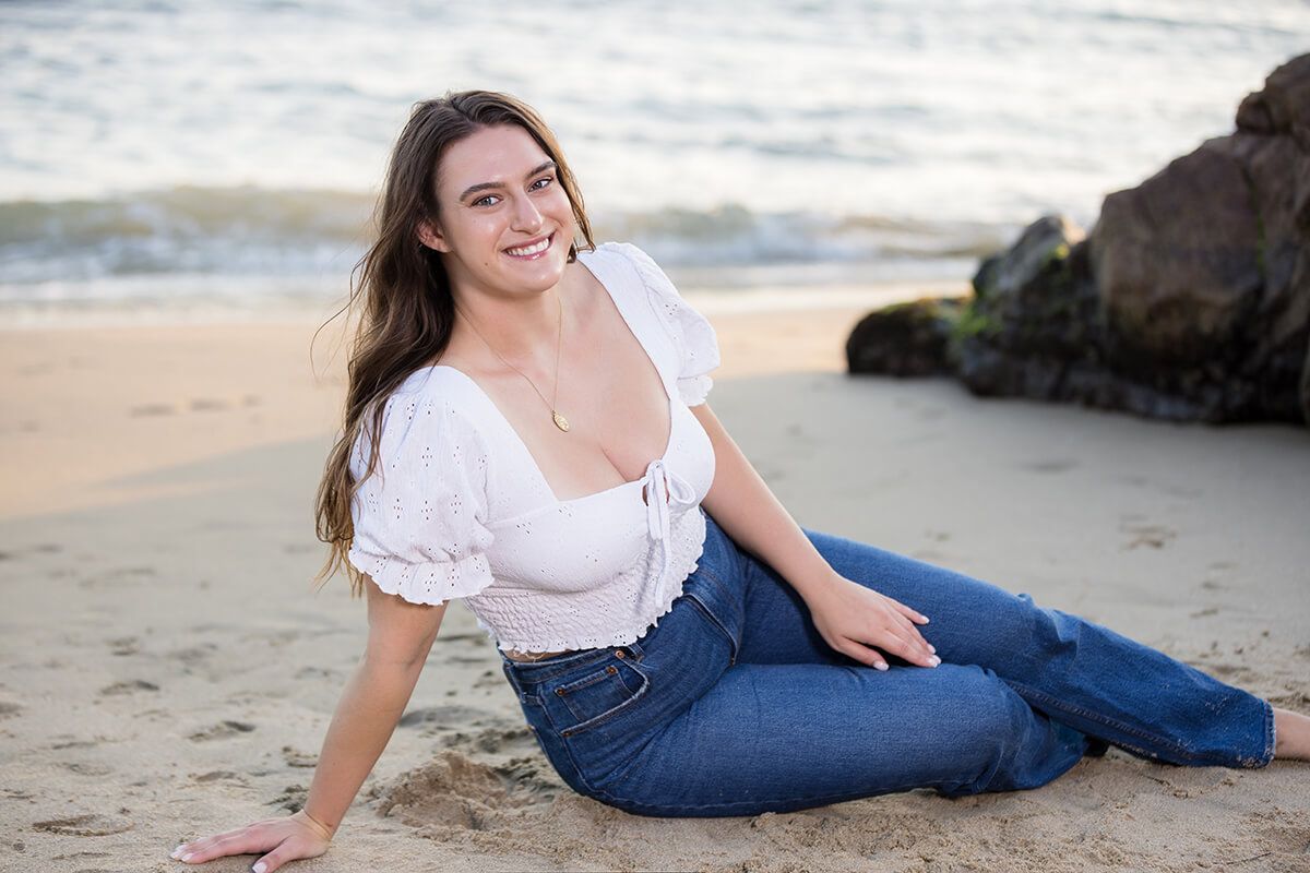 Natural light beach portrait of a young woman sitting on the sand at Picnic Beach in Laguna Beach, wearing a white eyelet blouse and denim jeans, photographed by Orange County senior and family photographer Blue Sky’s Studio.
