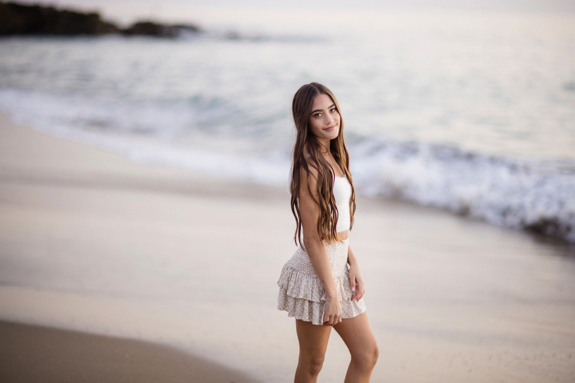A woman in a white dress is standing on a beach near the ocean.