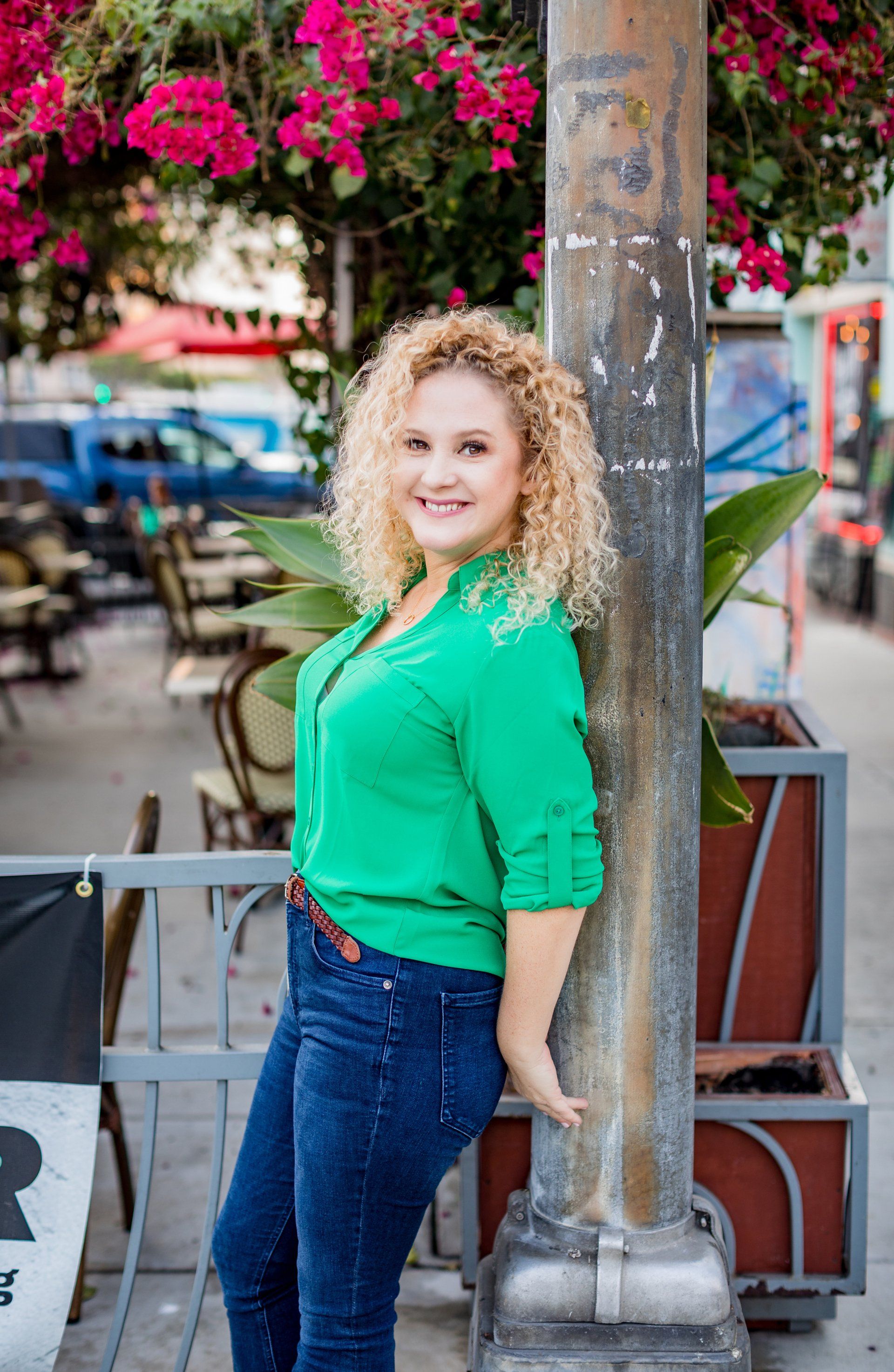 A woman in a green shirt and blue jeans is leaning against a pole.