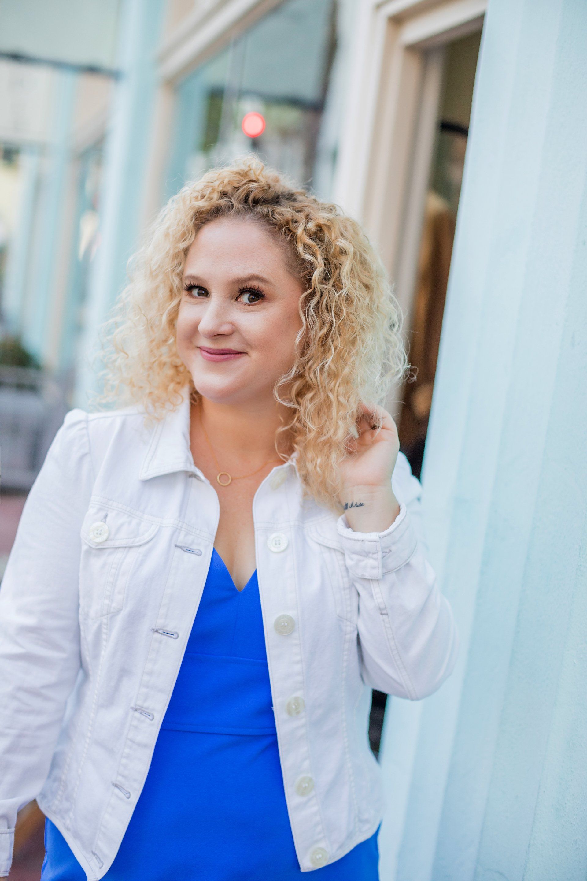 A woman wearing a blue dress and a white jacket is standing in front of a building.