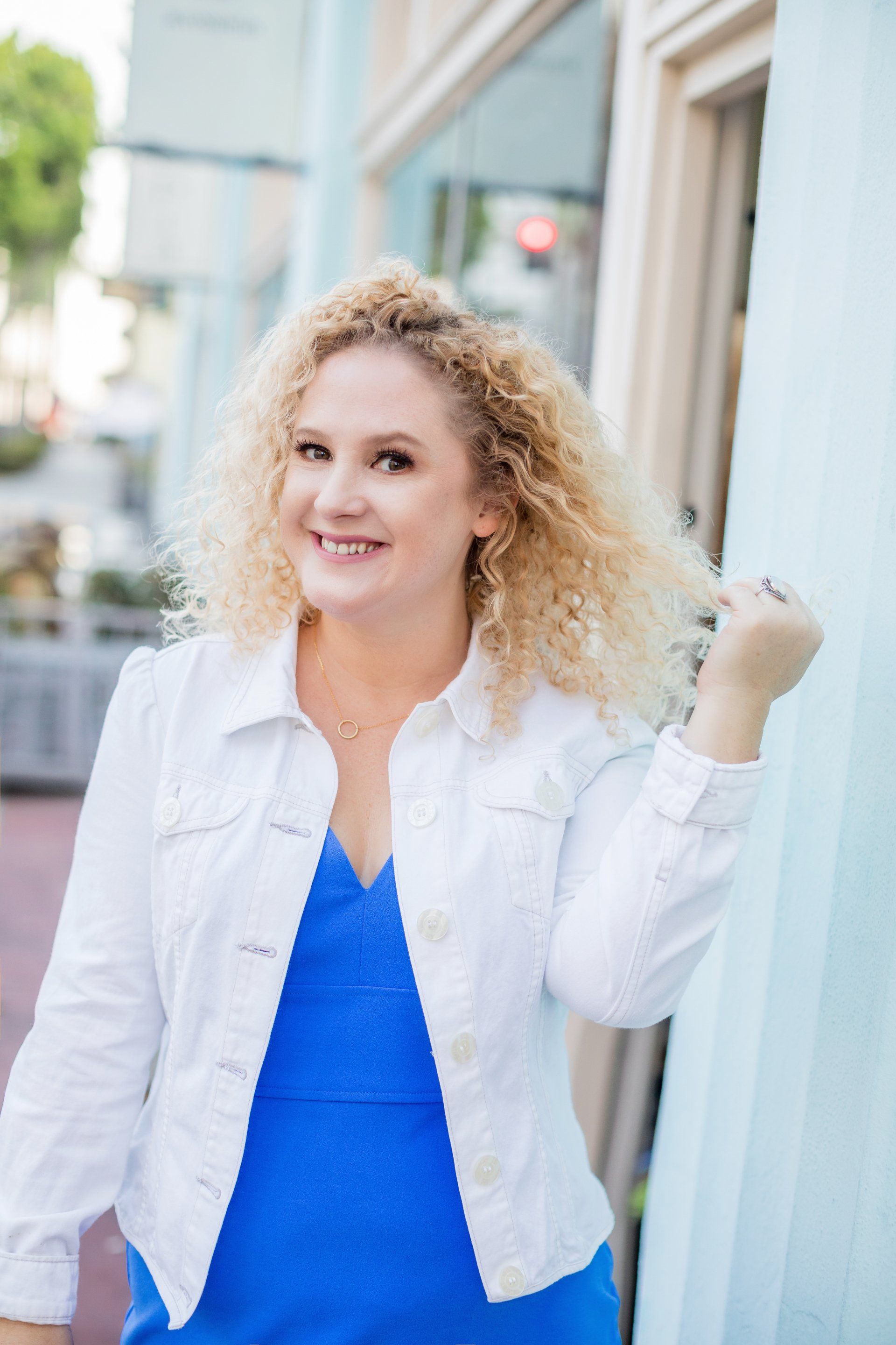 A woman wearing a blue dress and a white jacket is standing in front of a building.