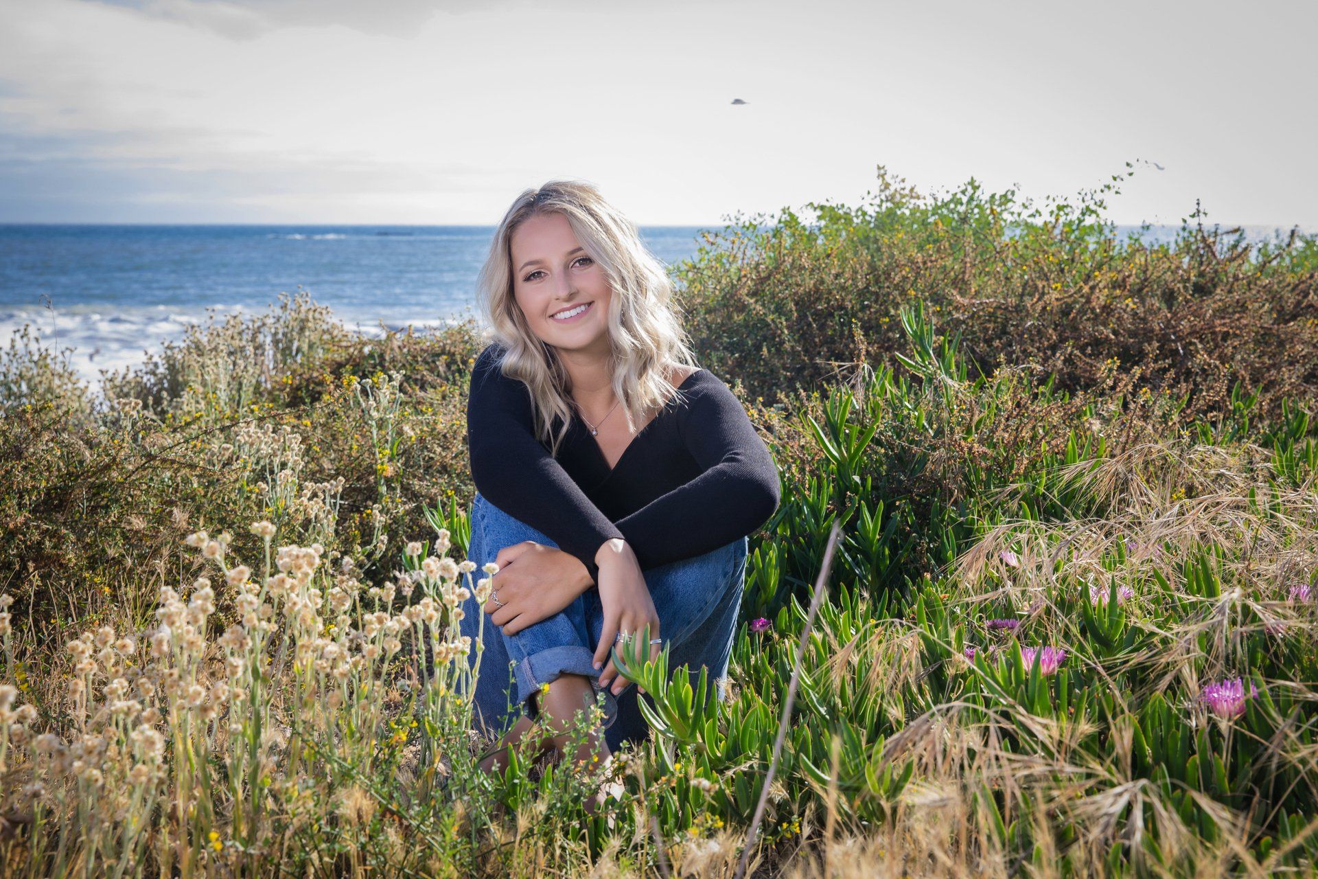 A woman is sitting in a field of flowers with the ocean in the background.