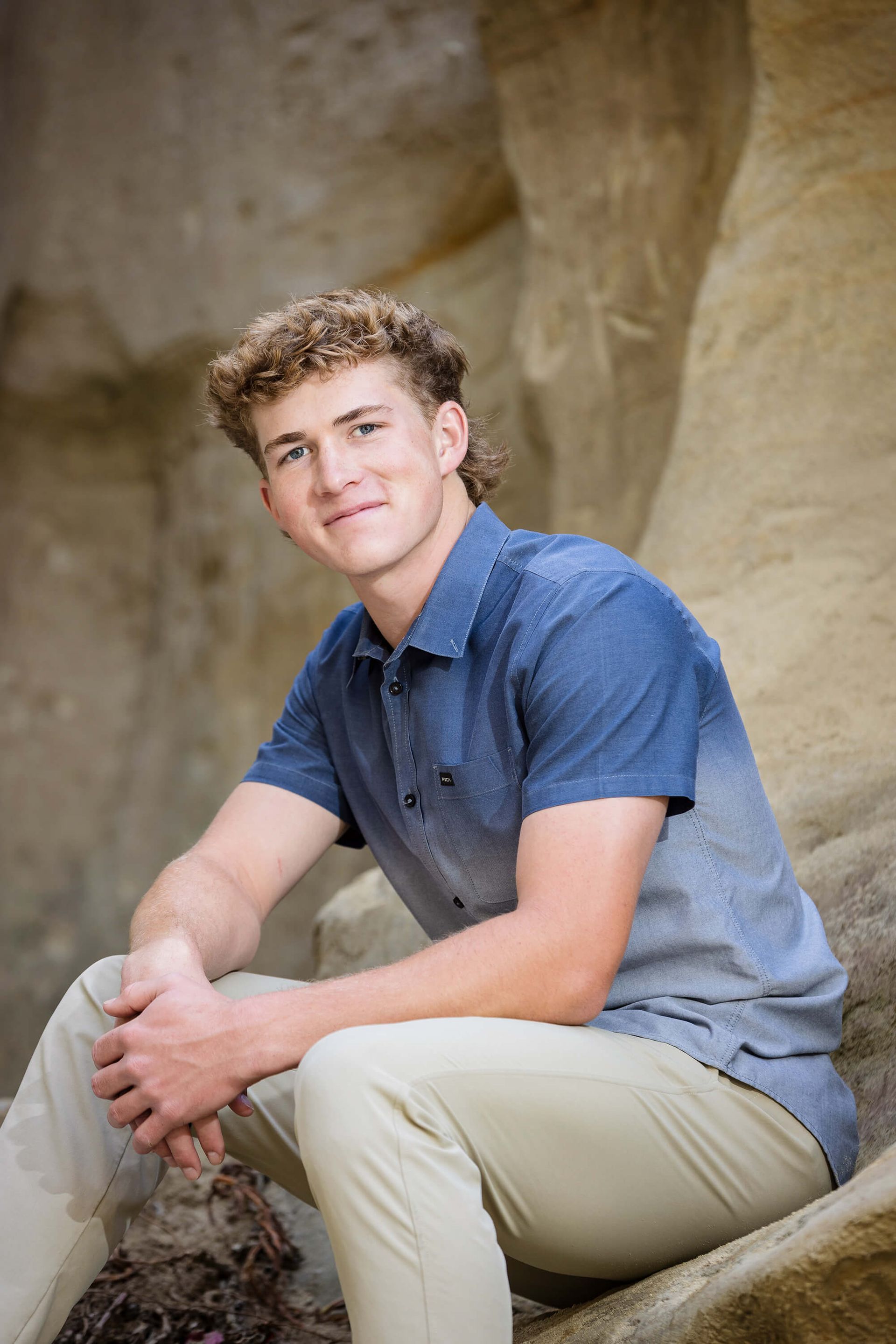 Orange County senior boy sitting on sandstone rock during his high school portrait session in Laguna Beach, wearing a blue button-down shirt and khaki pants — captured by Blue Sky’s Studio