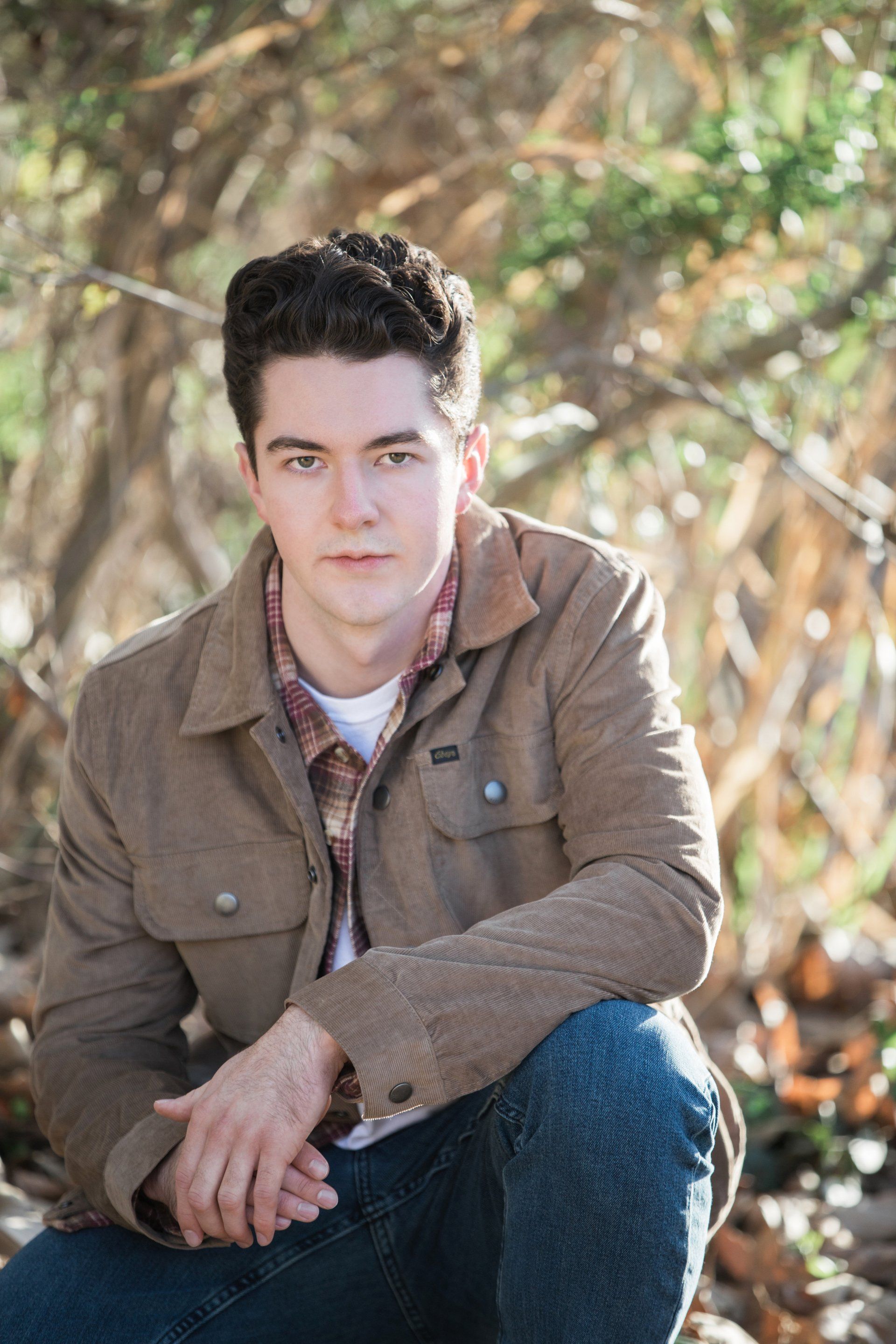 A young man in a brown jacket is sitting on the ground with his hands folded.