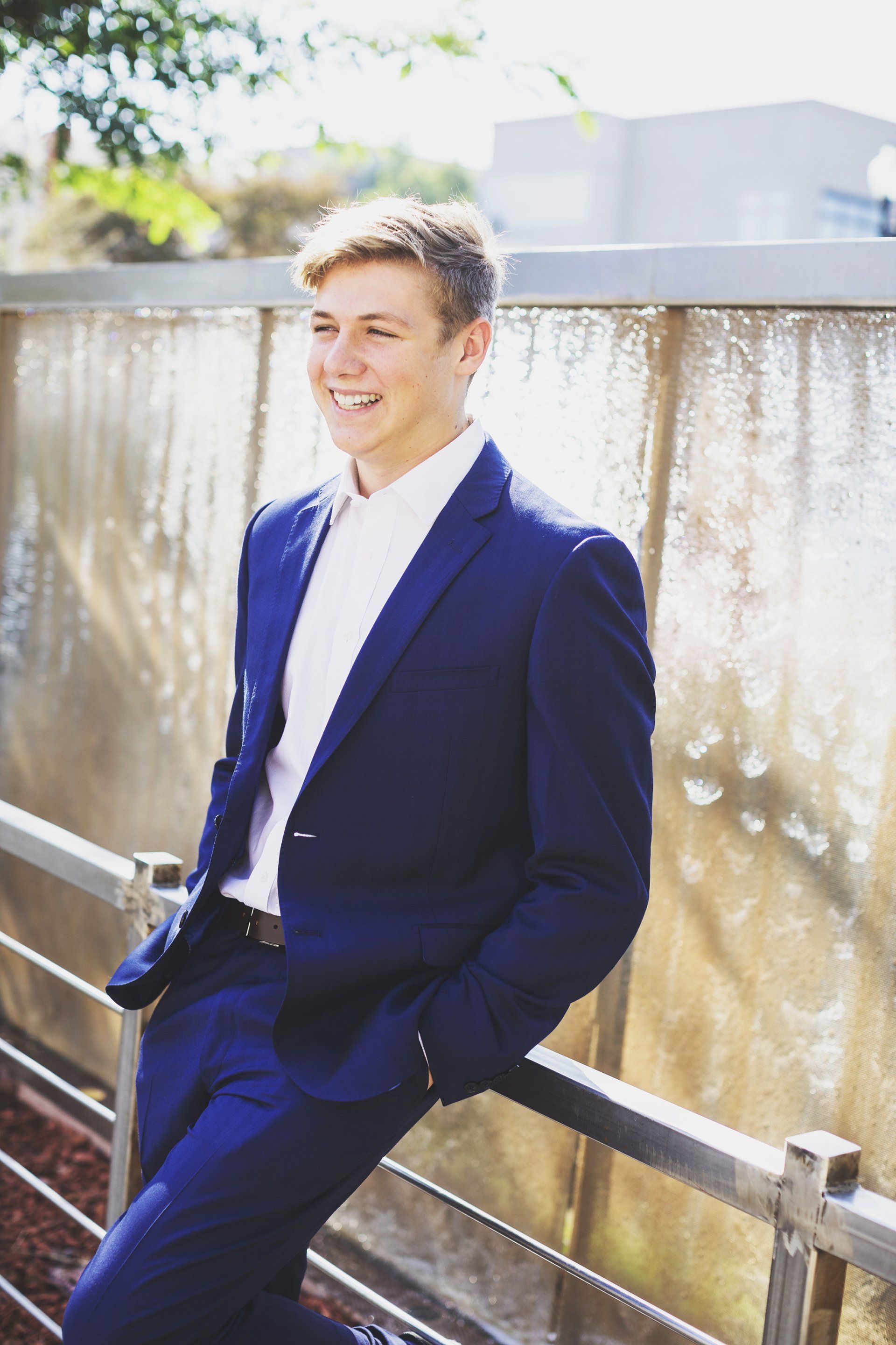 A young man in a blue suit and white shirt is leaning against a railing.