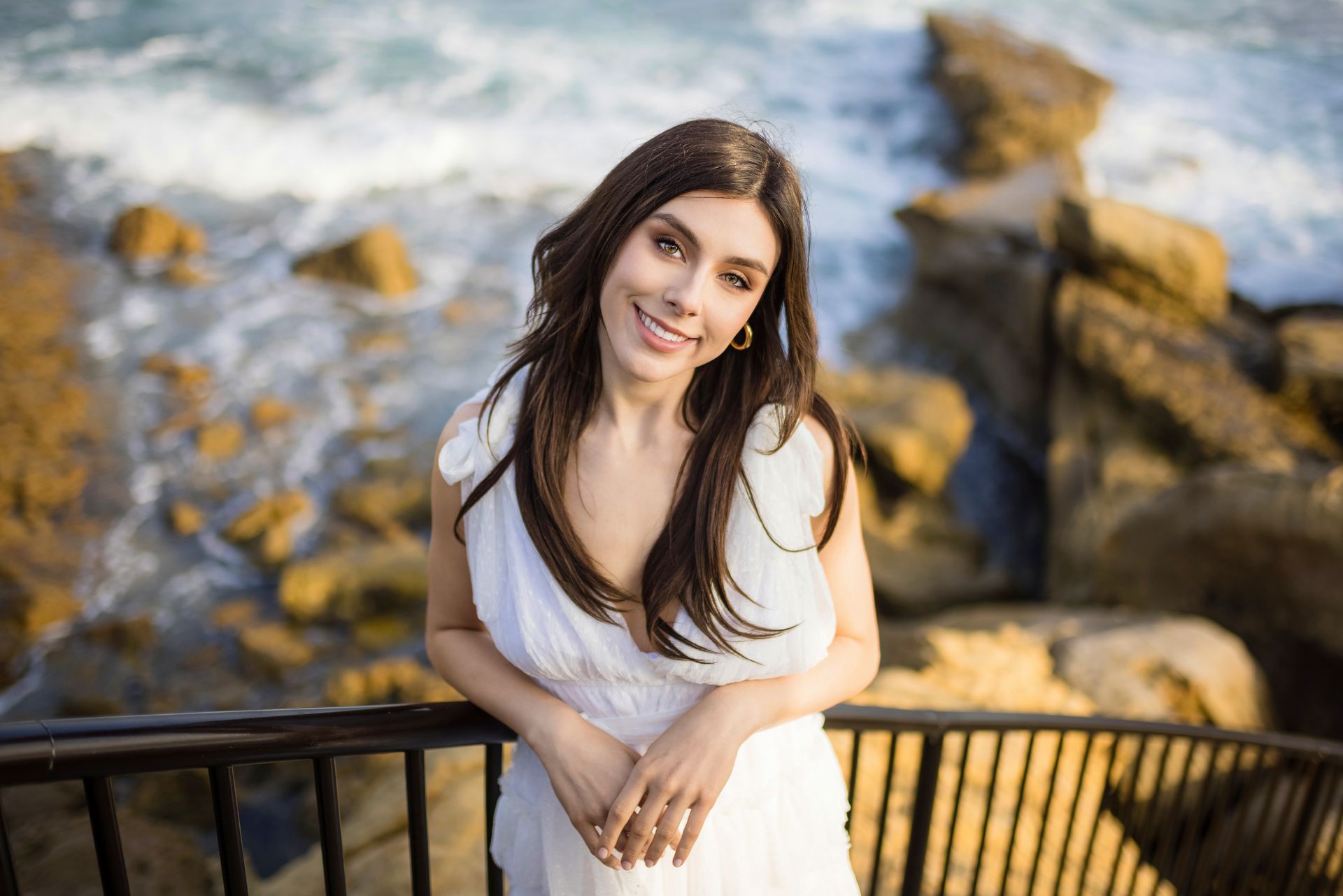 A woman in a white dress is leaning on a railing near the ocean.