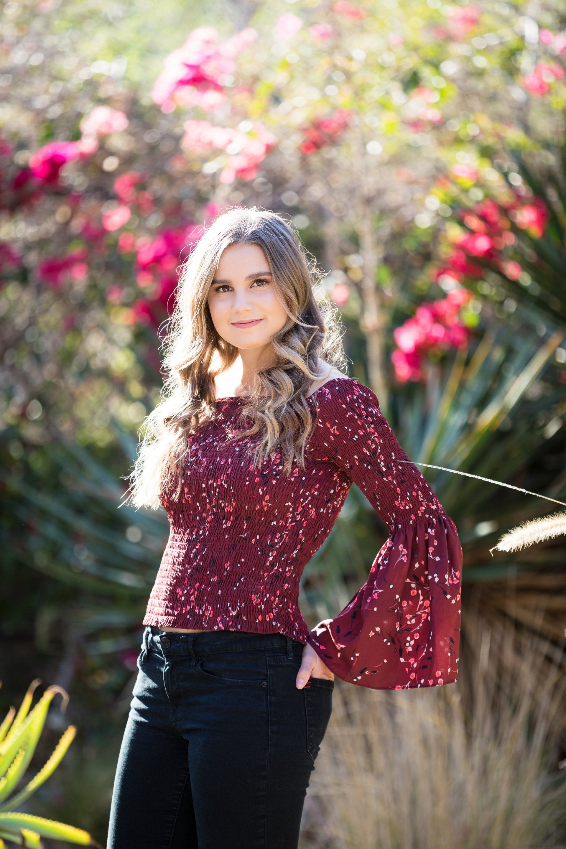 A young woman is standing in front of a bush with pink flowers.
