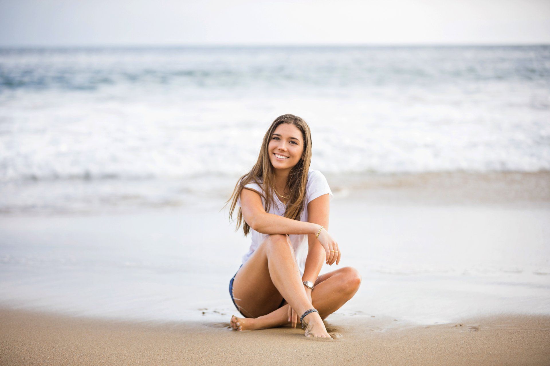 A woman is sitting on the beach with her legs crossed and smiling.