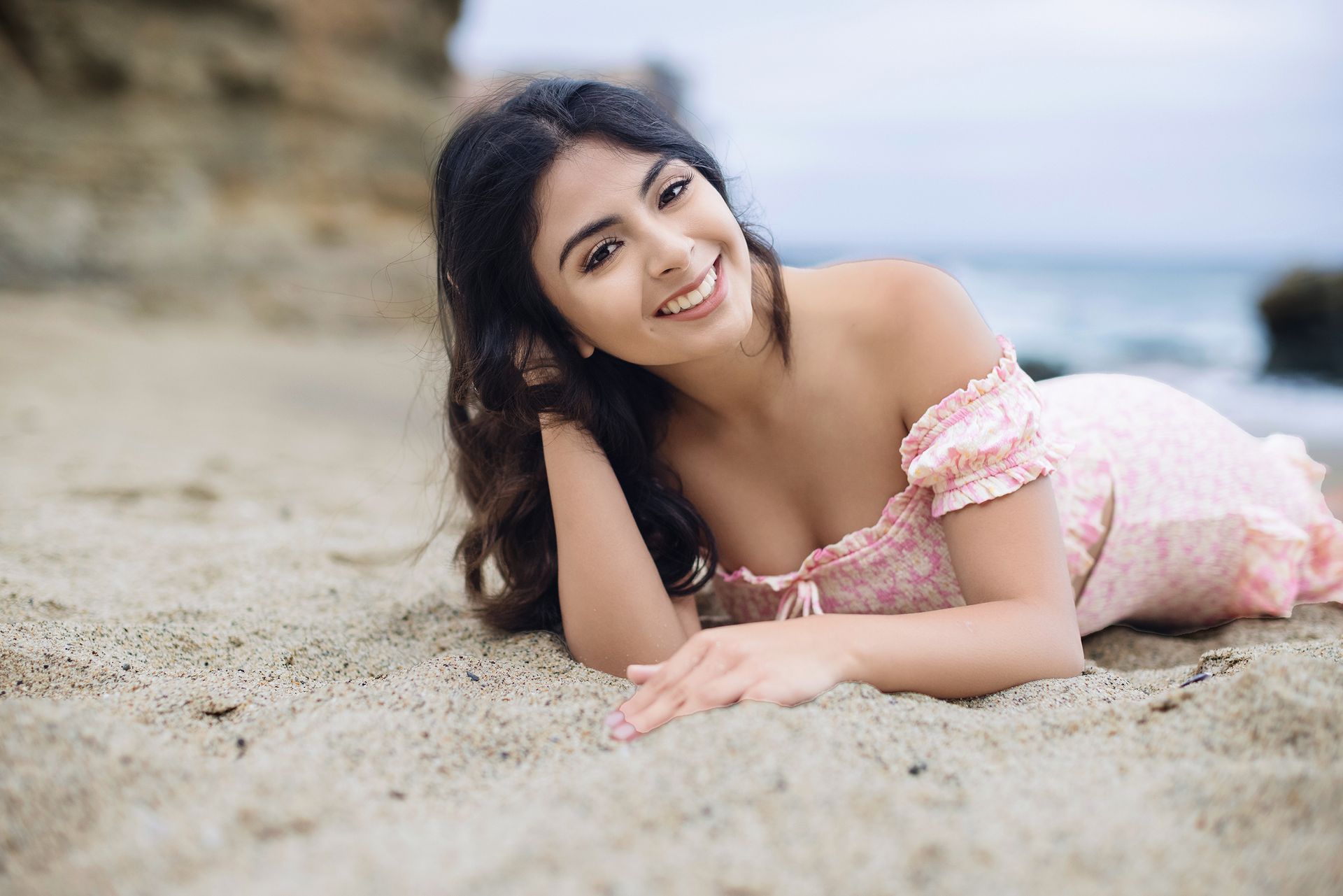 A woman in a pink dress is laying on the beach and smiling.