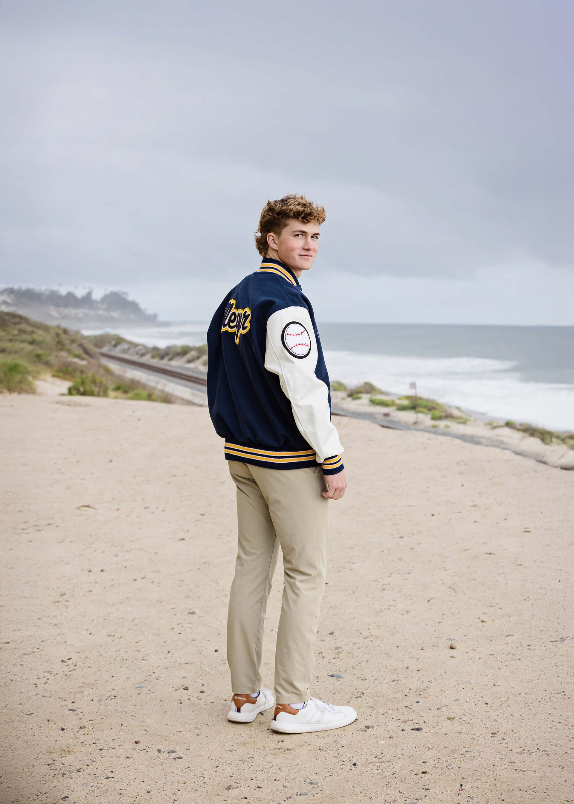 Orange County high school senior wearing a varsity letterman jacket during his senior portrait session on the beach in Laguna Beach, captured by Blue Sky’s Studio.