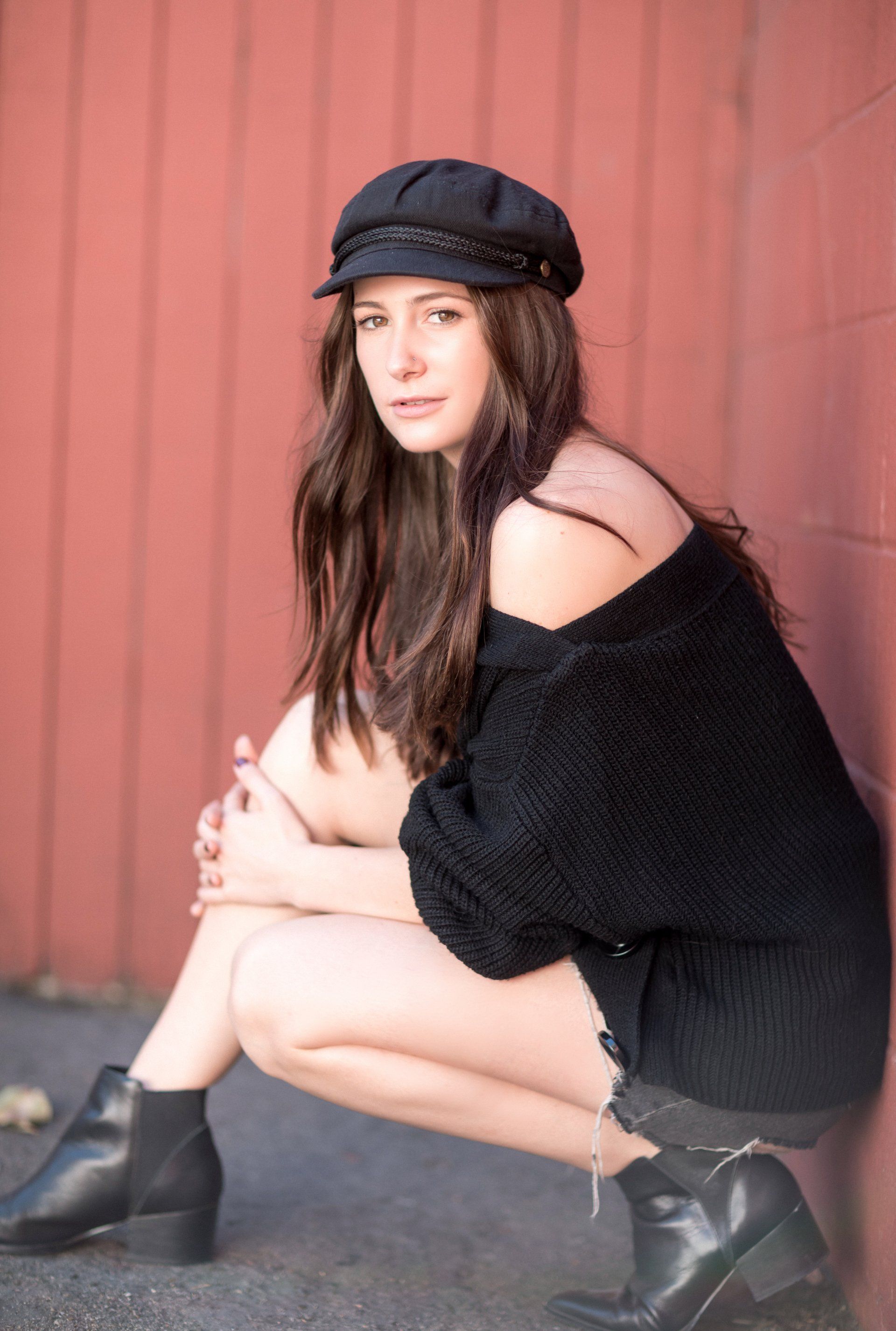 A young woman crouches against a red wall, wearing a black off-the-shoulder sweate