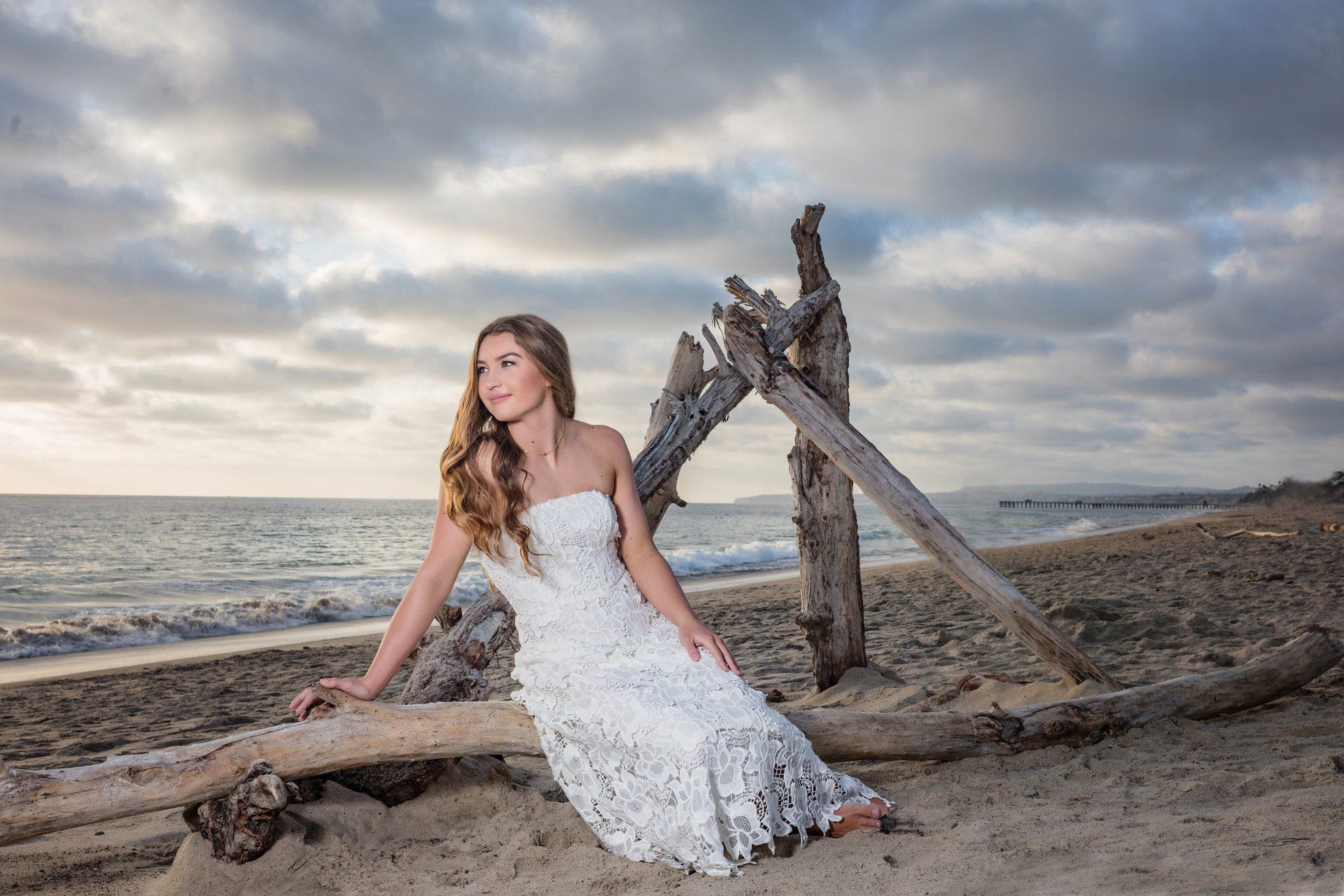 A woman in a white dress is sitting on a log on the beach.