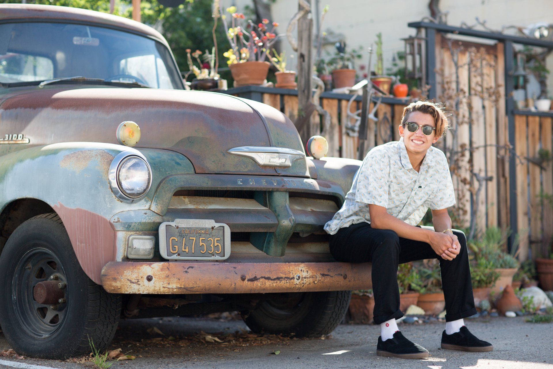 A young man is sitting on the front of an old rusty truck.
