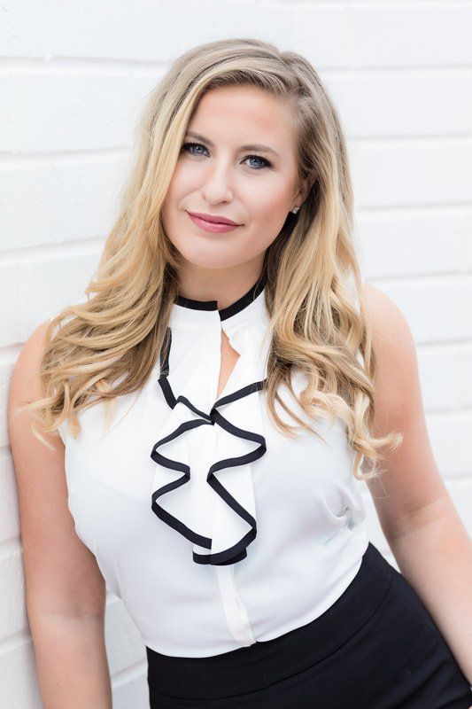A woman in a white top and black skirt is standing in front of a white wall.
