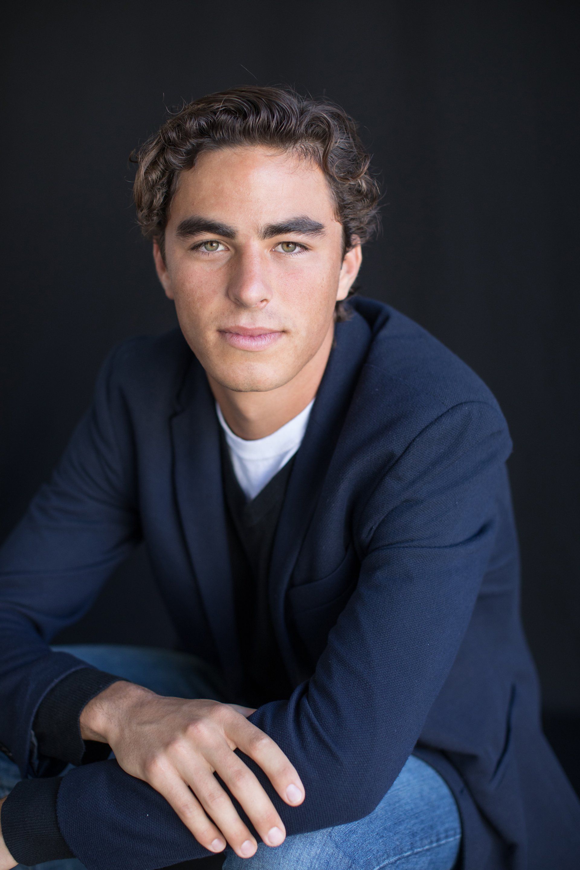 A young man in a blue jacket is sitting down with his legs crossed.