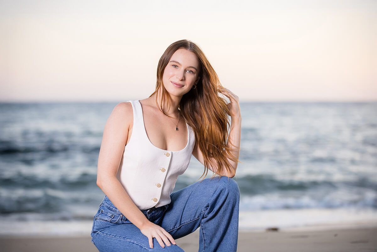 Sunset beach portrait of a young woman with long red hair in jeans and a white tank top, captured in Laguna Beach by Orange County family and senior portrait photographer Blue Sky’s Studio.