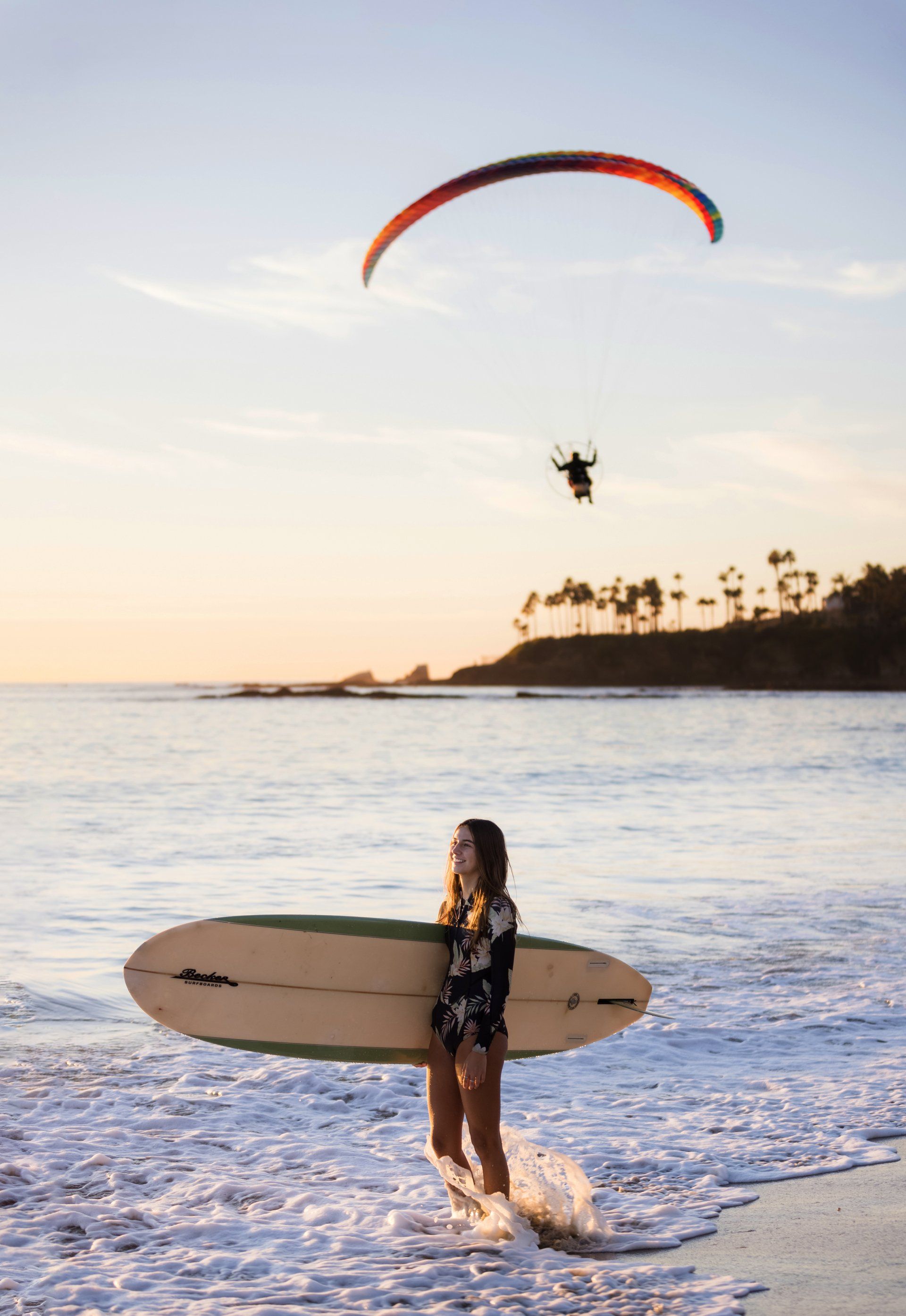 A woman is holding a surfboard on the beach while a paraglider flies overhead.