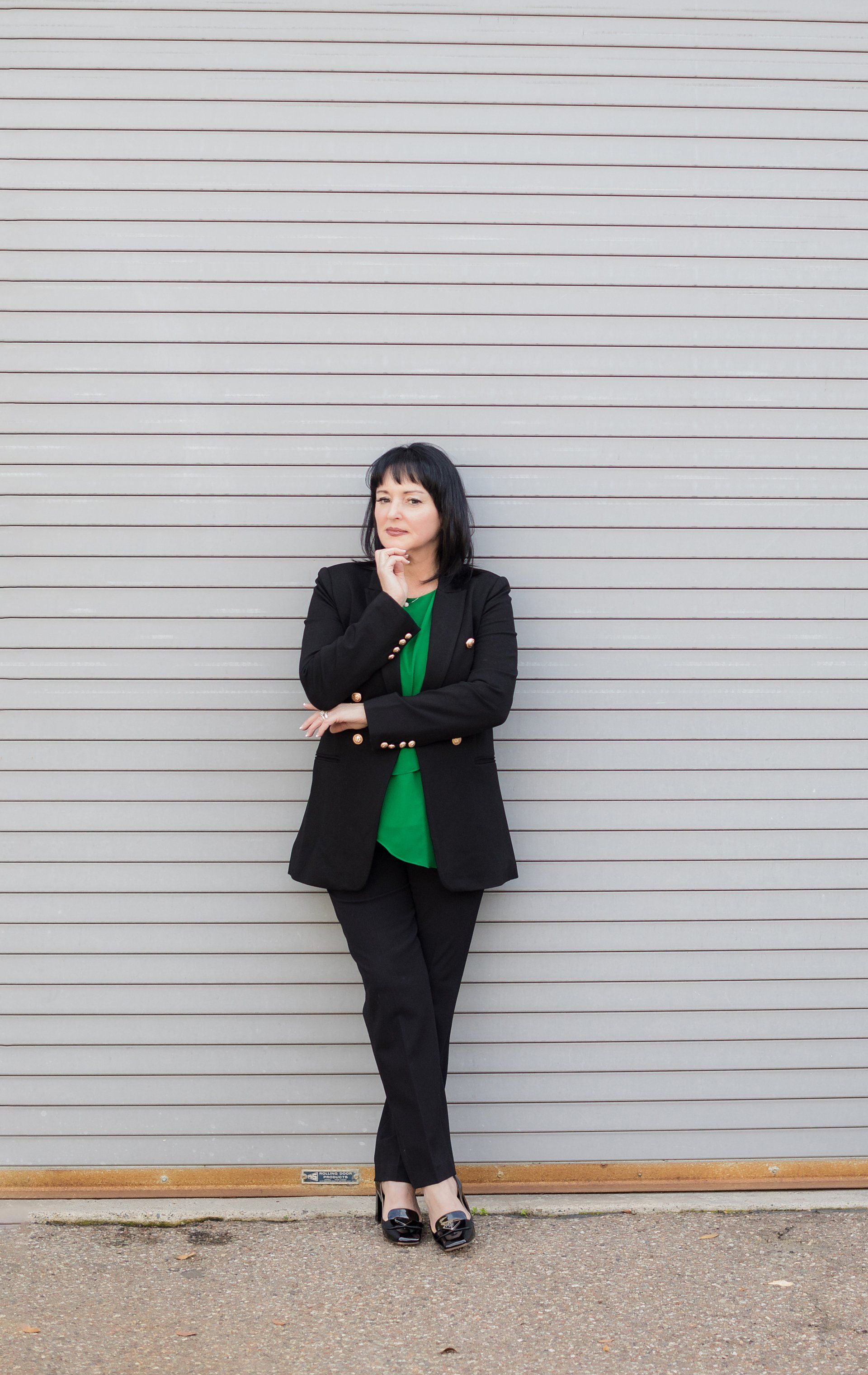 A woman in a black suit and green shirt is standing in front of a garage door.