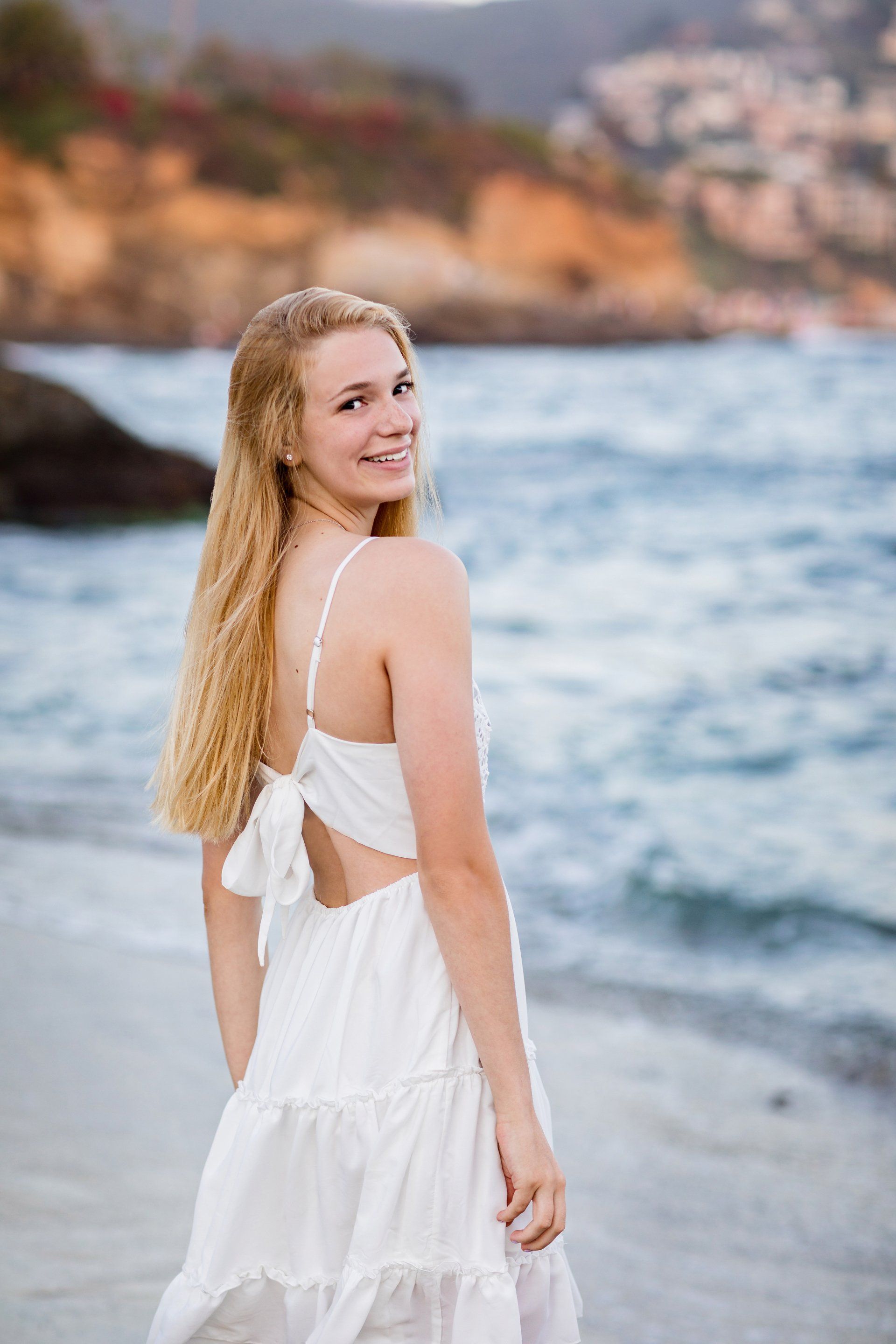 A woman in a white dress is standing on a beach looking at the ocean.