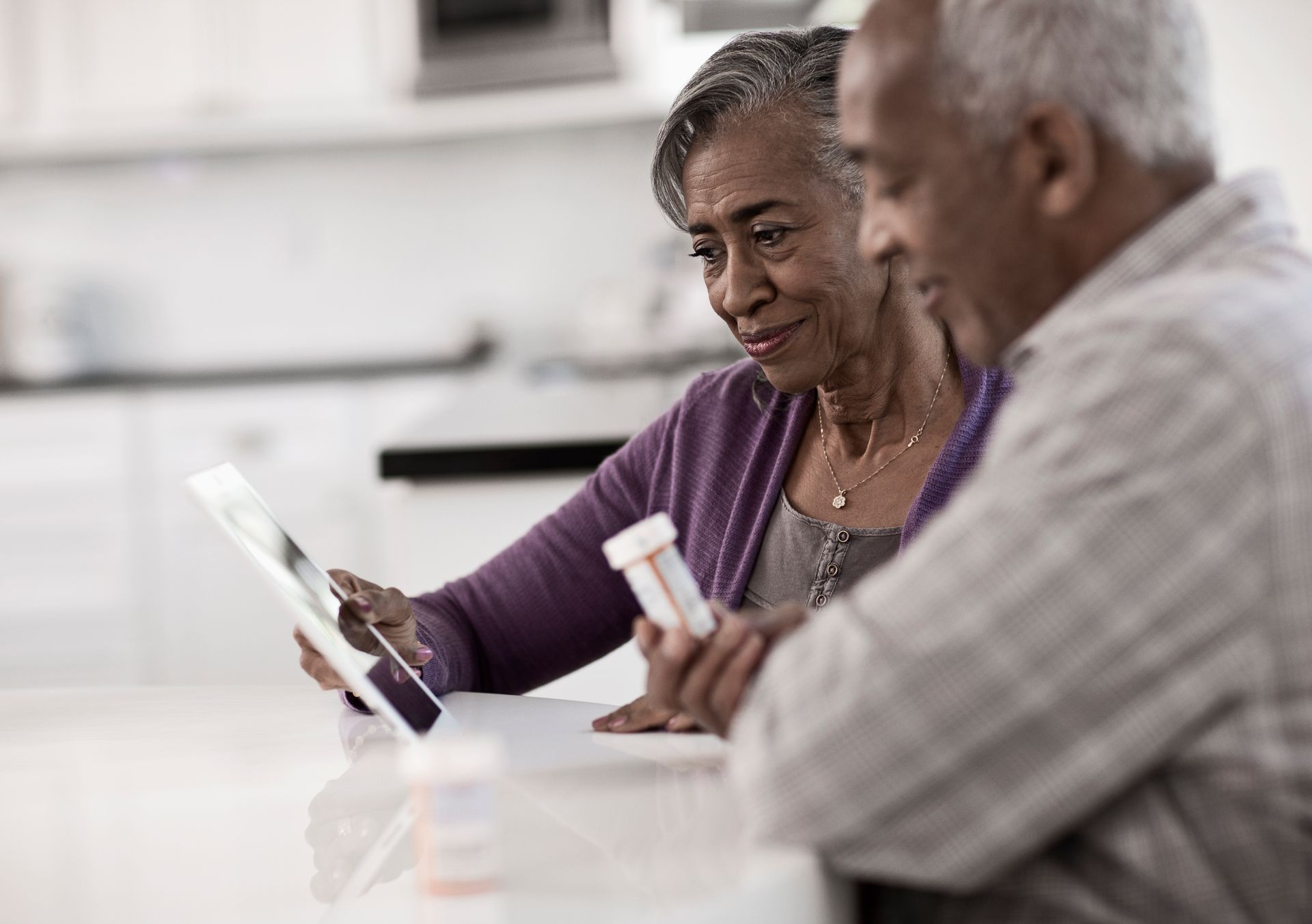 Older couple looking at a tablet, examining pills in a kitchen, smiling.