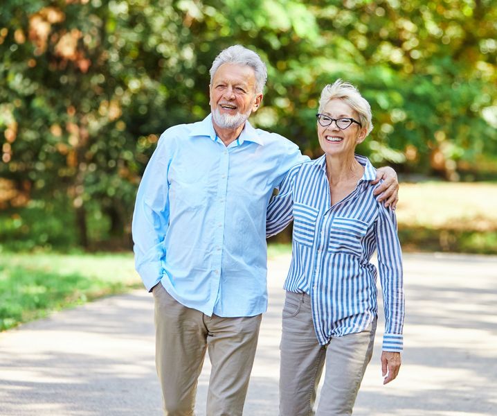 Elderly couple walking on path, man has arm around woman. Green trees in background.