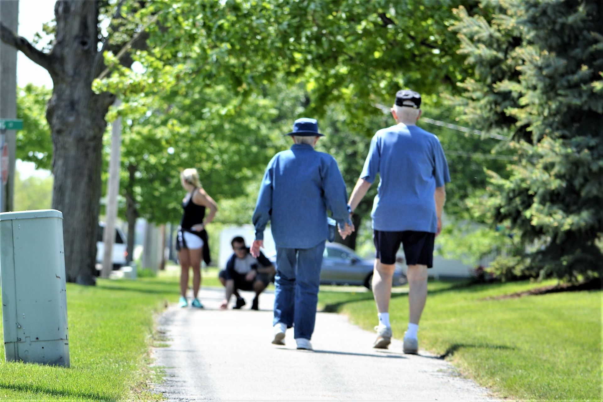 Two men holding hands walk on a path; others are nearby. Sunny day.