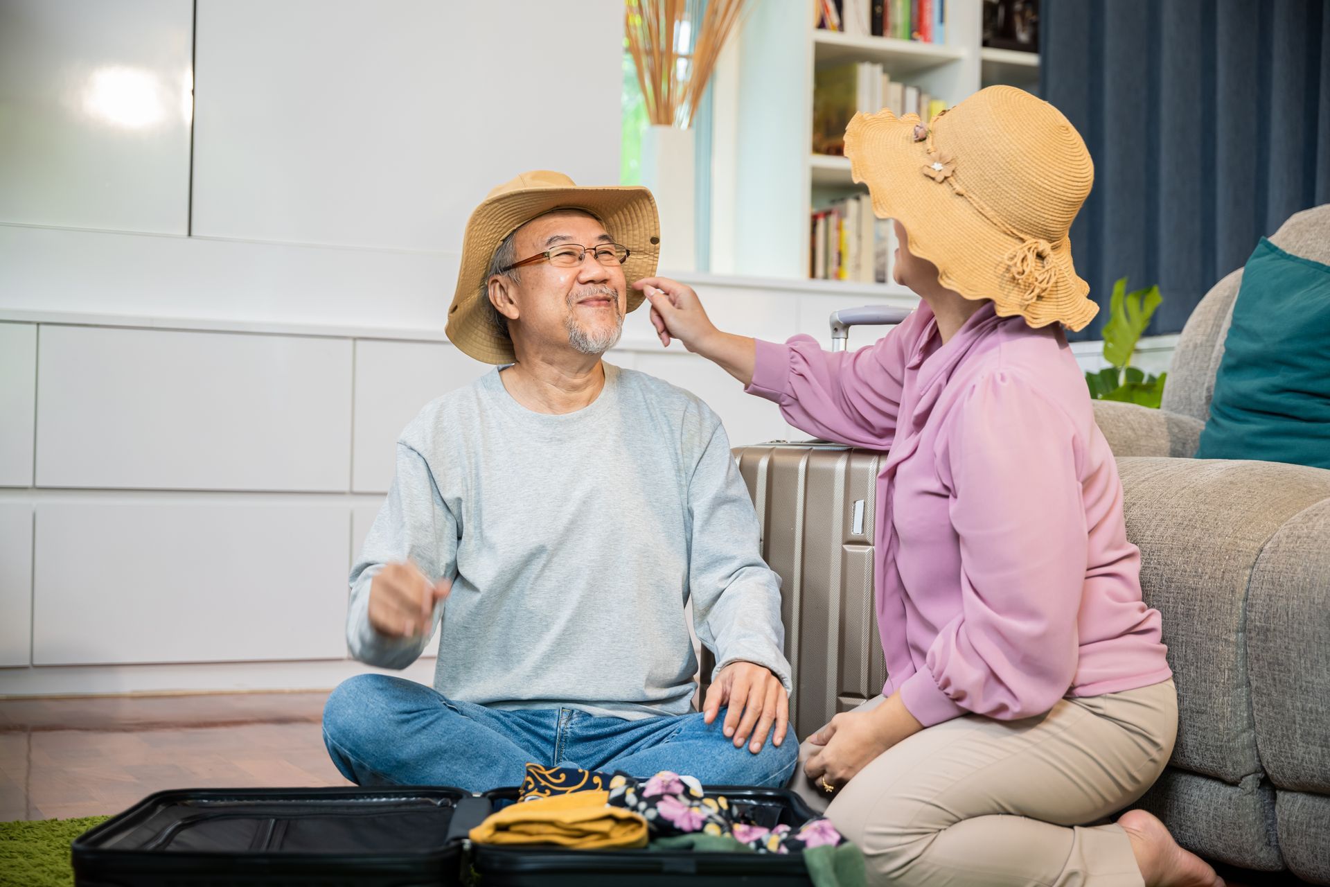 An older Asian couple packing suitcases, preparing for a trip at home. Woman adjusting man’s straw hat.