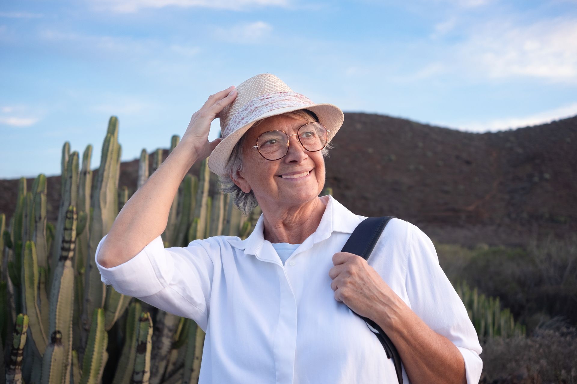 Woman in white shirt and straw hat, smiling, touching hat, outdoors with cactus and hills.