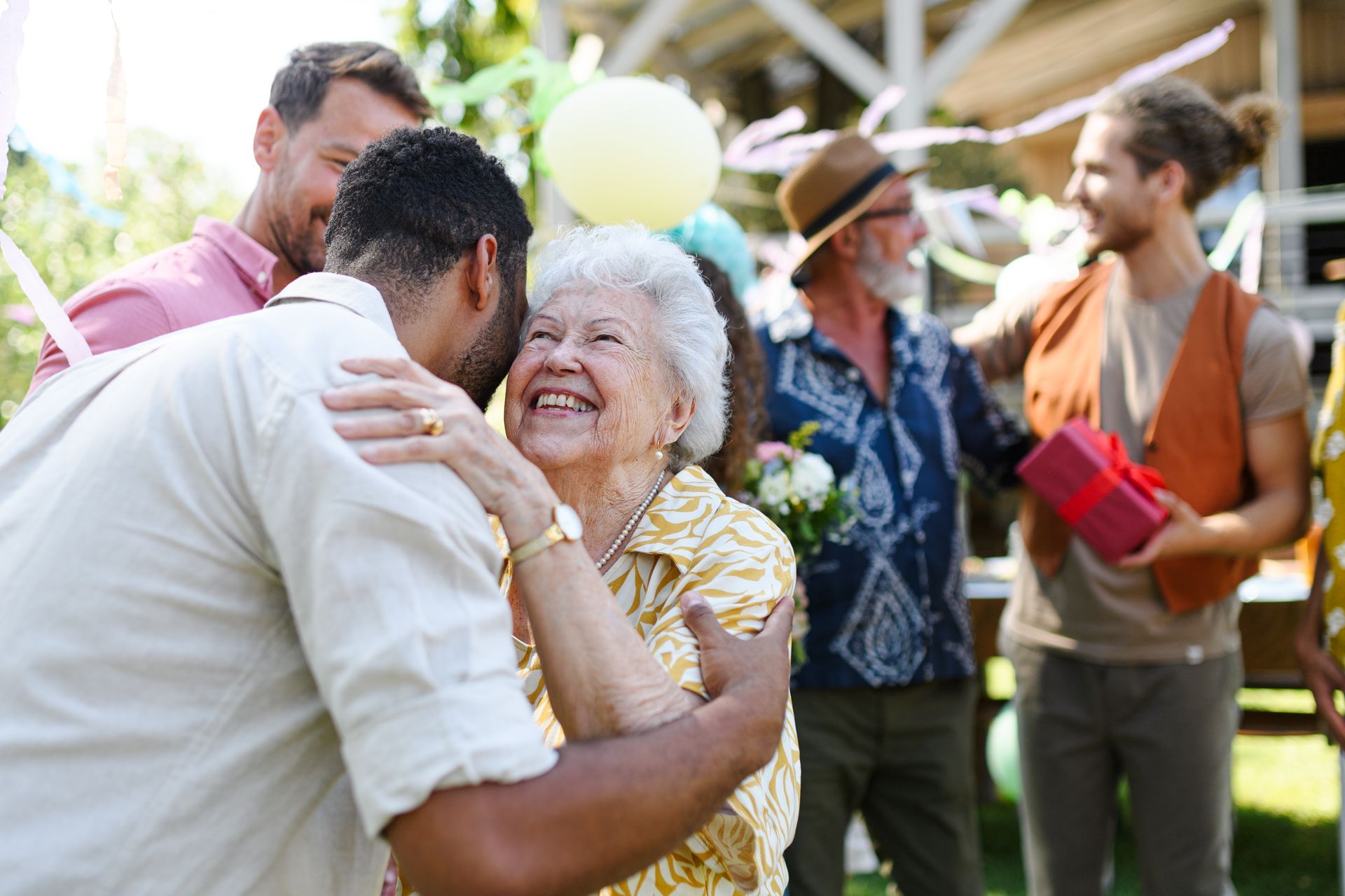 A joyful group hugs a smiling elderly woman at an outdoor celebration with gifts and balloons.