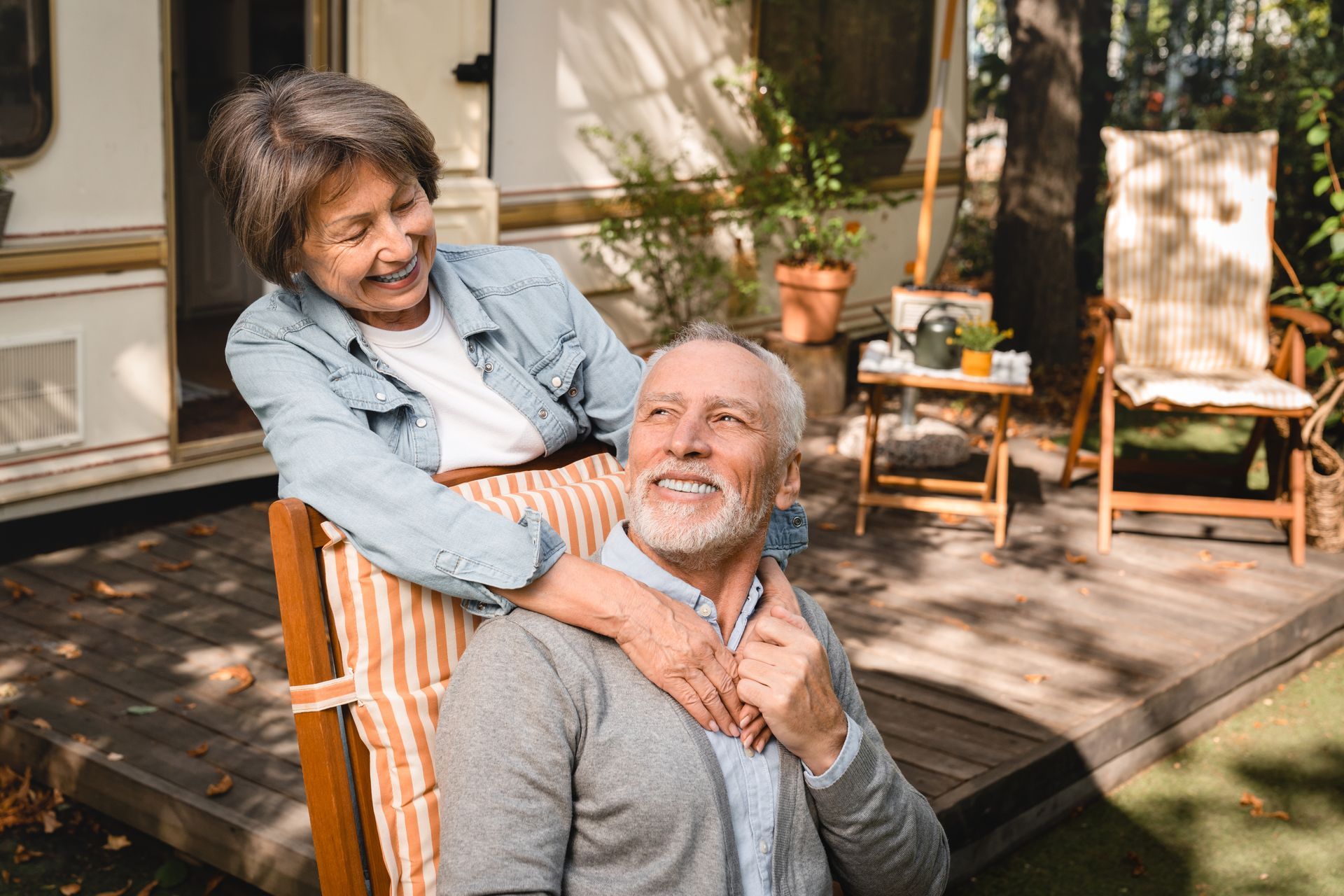 Smiling elderly couple outdoors; woman with arms around man's shoulders. Camper in background.