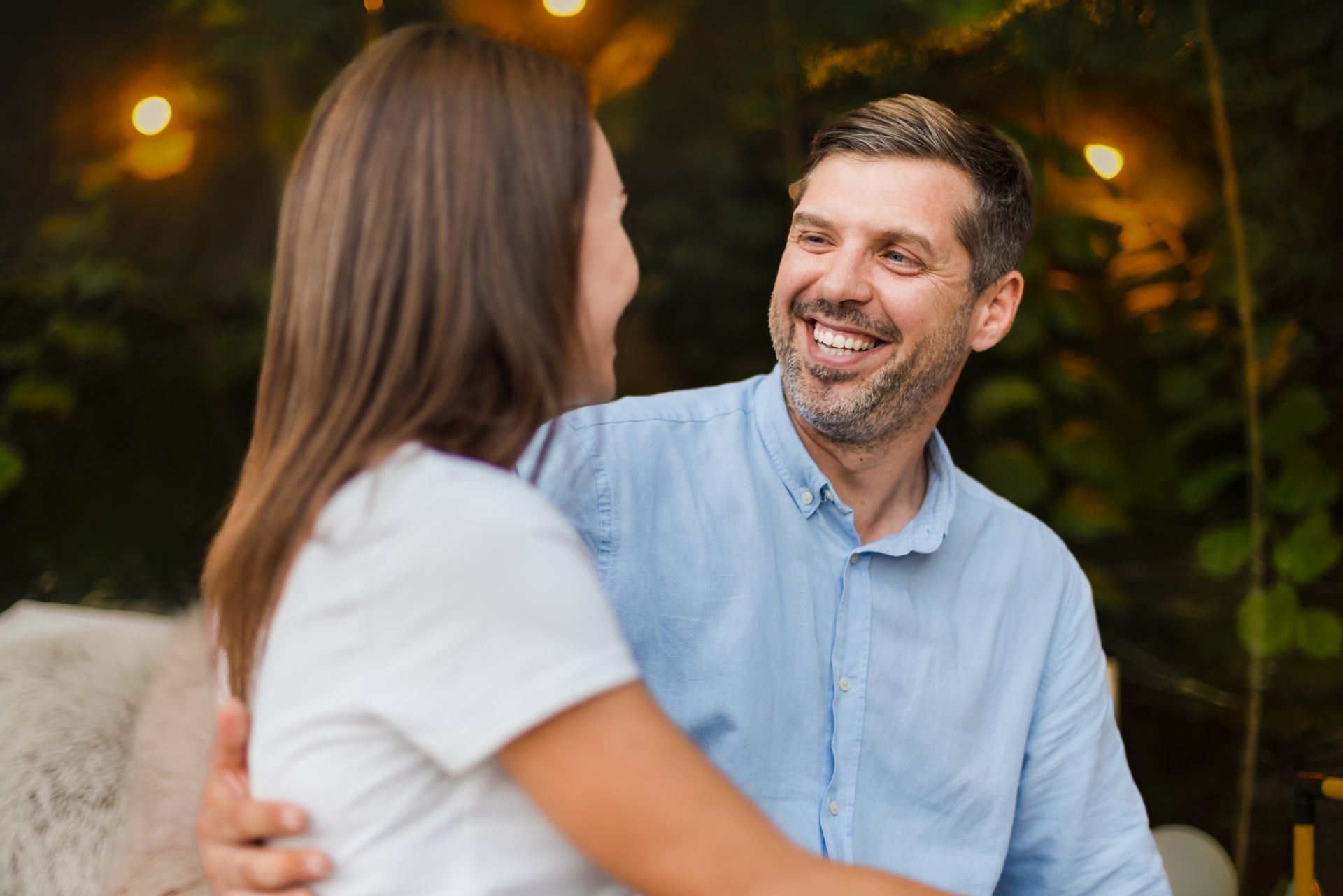 Man with his arm around a woman, smiling at her outdoors under fairy lights.