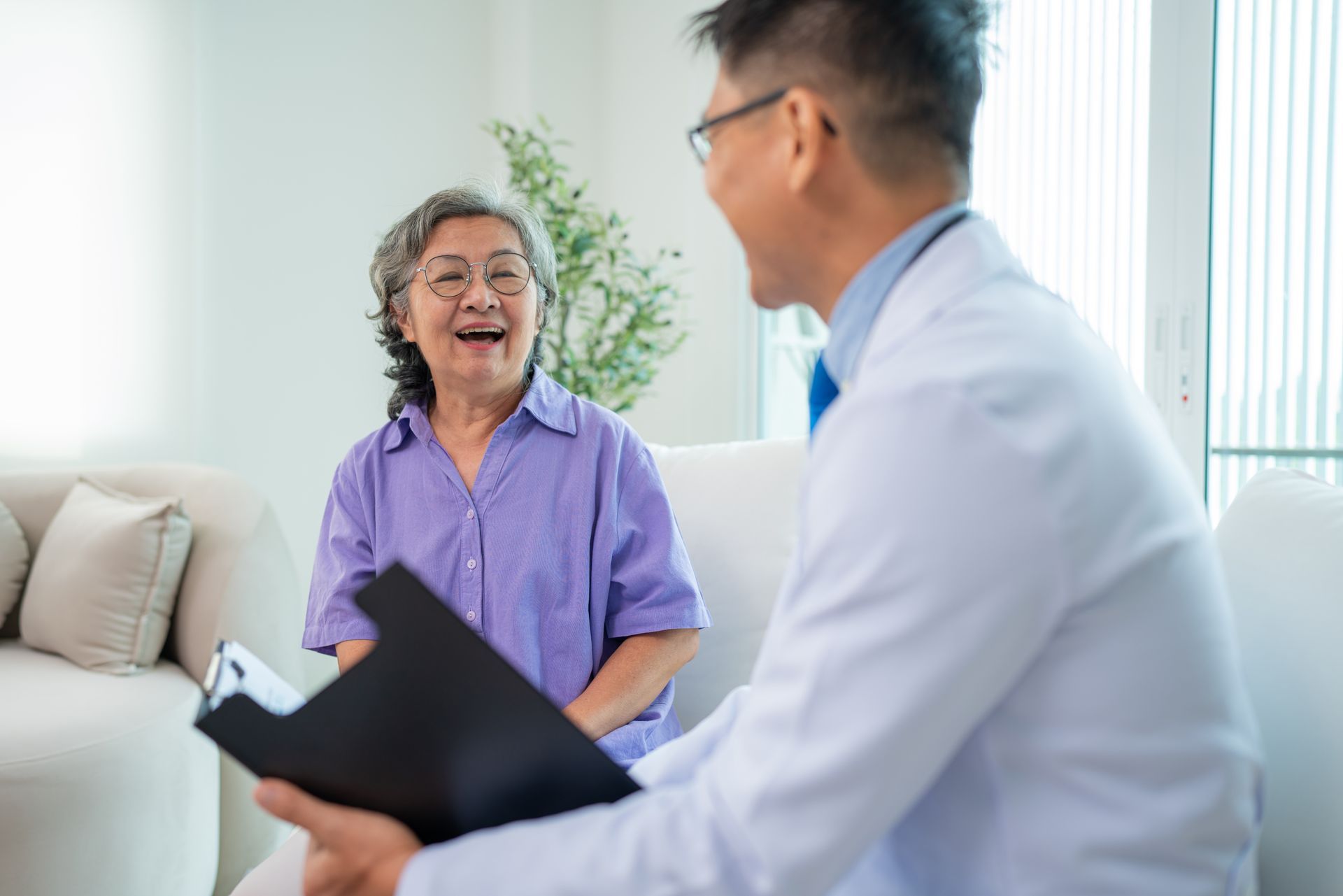 Doctor in white coat smiles at an elderly woman, holding a clipboard in a living room setting.