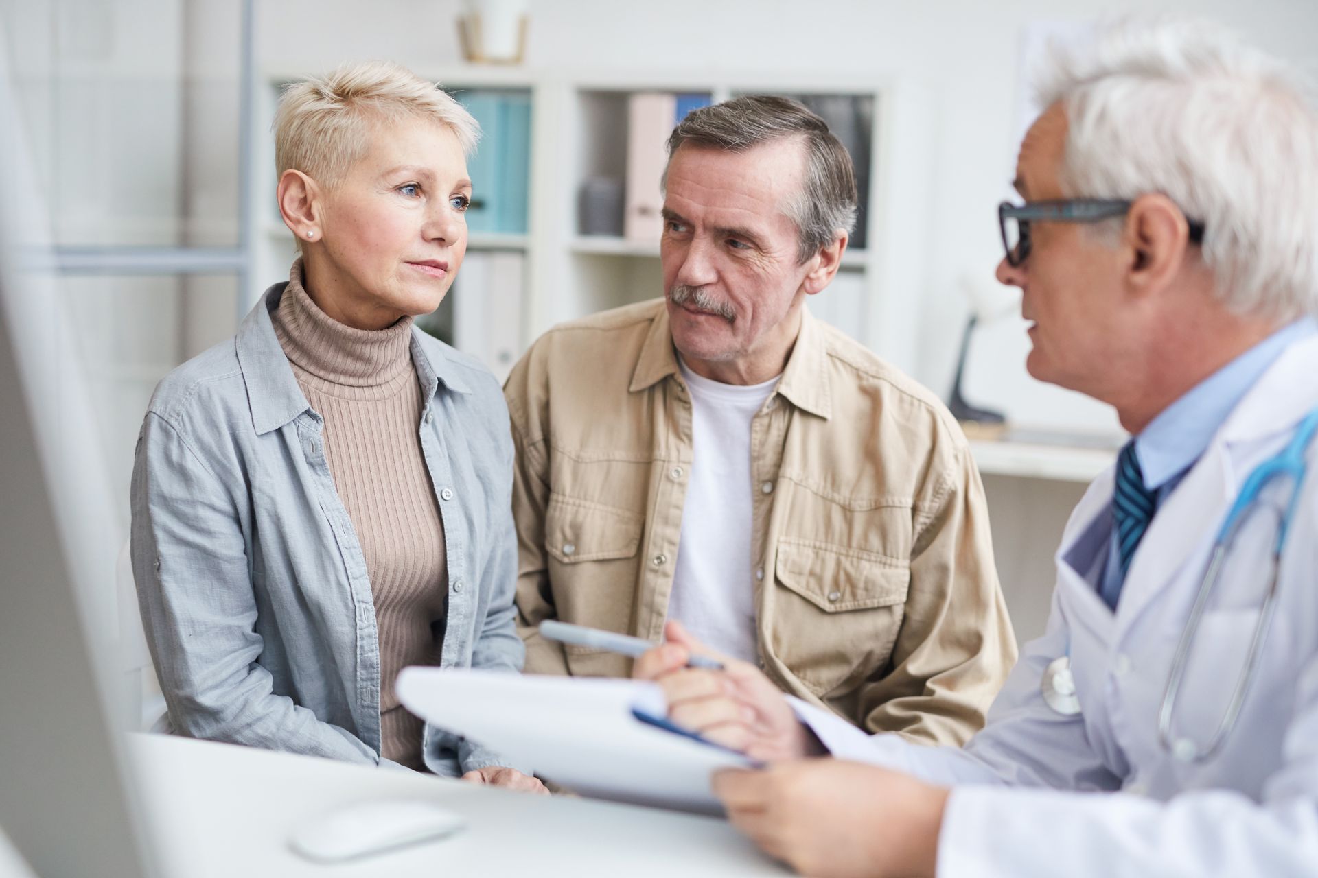 A doctor in a white coat consults with an older couple in an office setting, looking at paperwork.