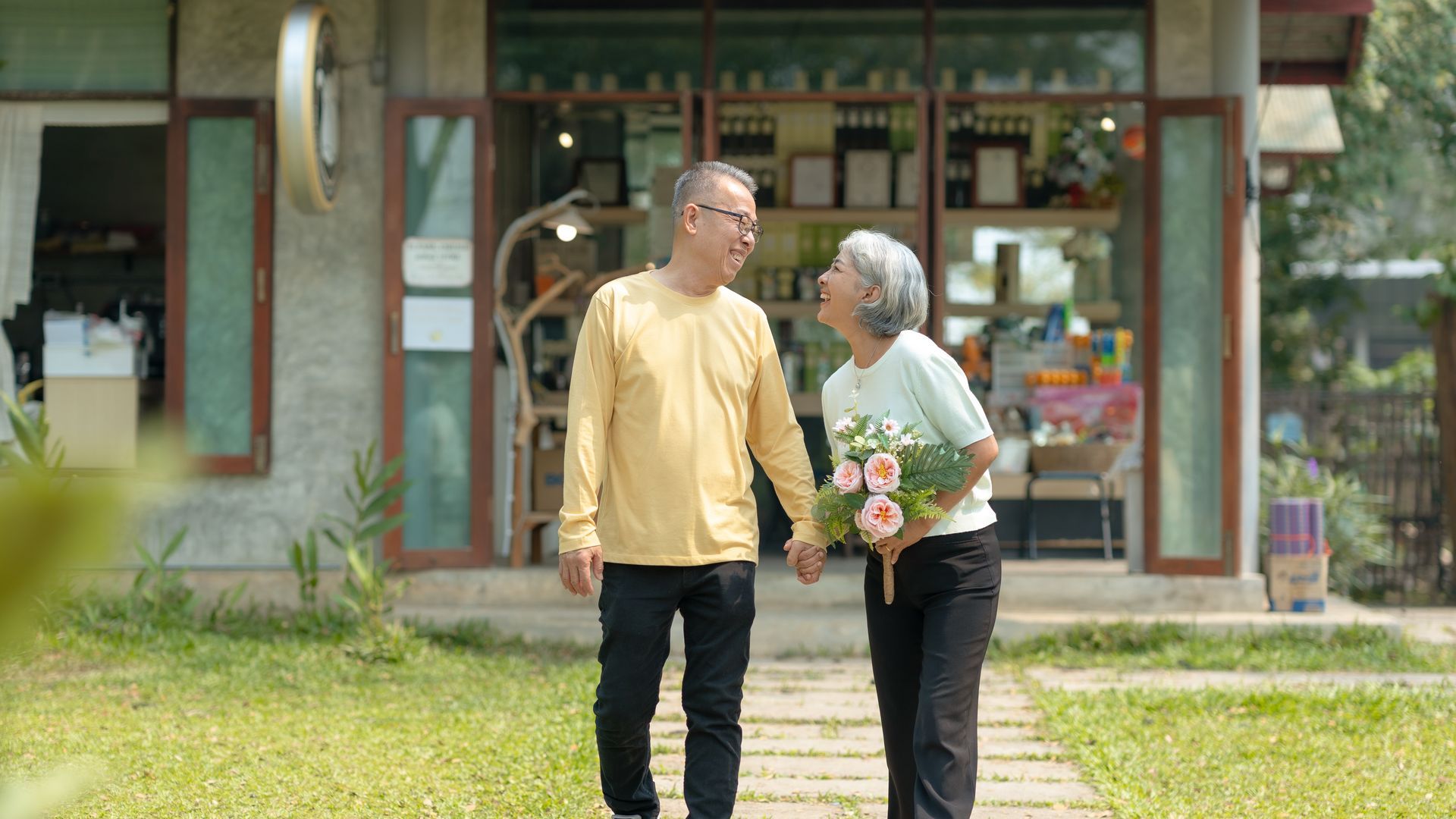 Elderly couple holding hands, walking from a flower shop, woman holds flowers, sunny day.