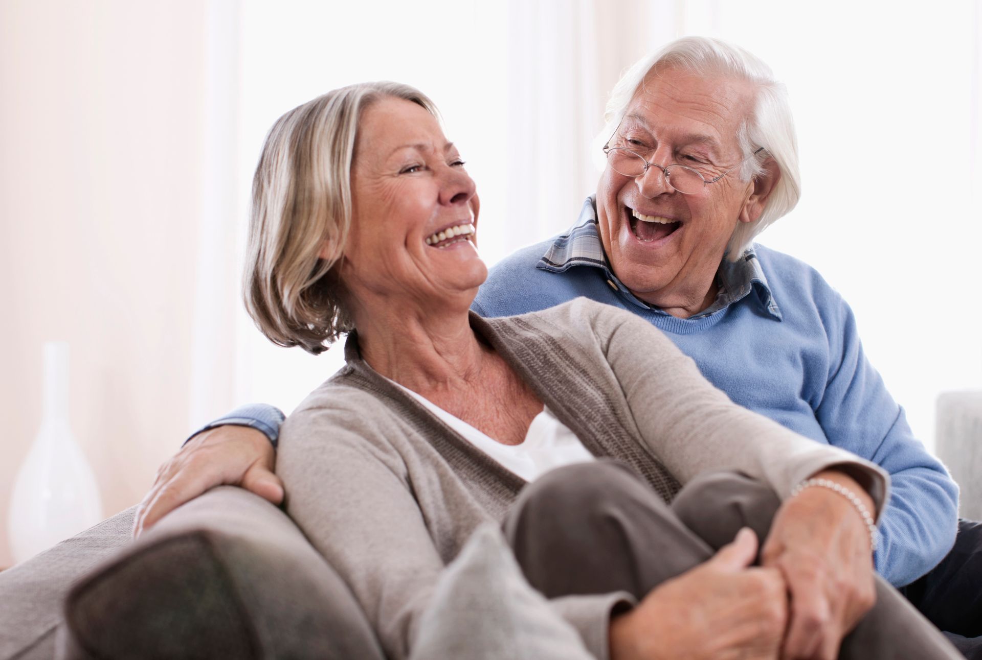 Smiling elderly couple on a couch, laughing together. Woman in cardigan, man in blue sweater.
