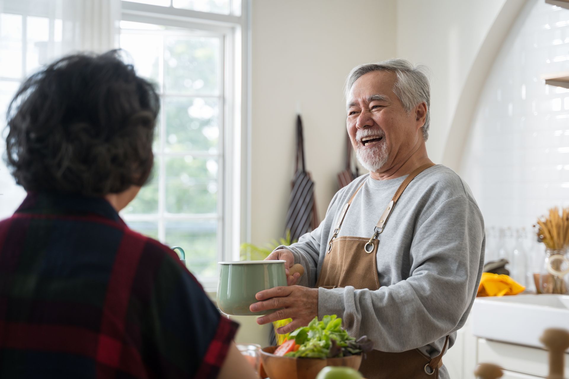 An elderly Asian man in a kitchen, smiling, holding a pot, and talking to someone out of frame.