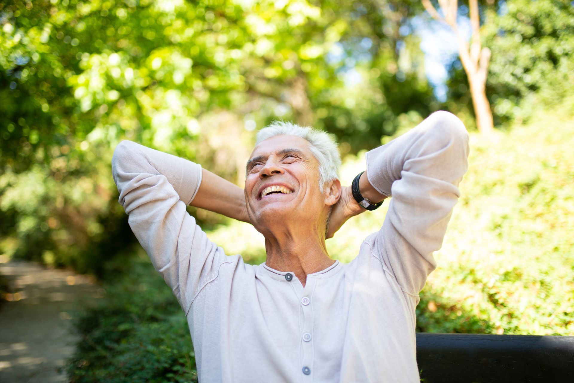 Smiling older man with hands behind head, outdoors with green trees in background.