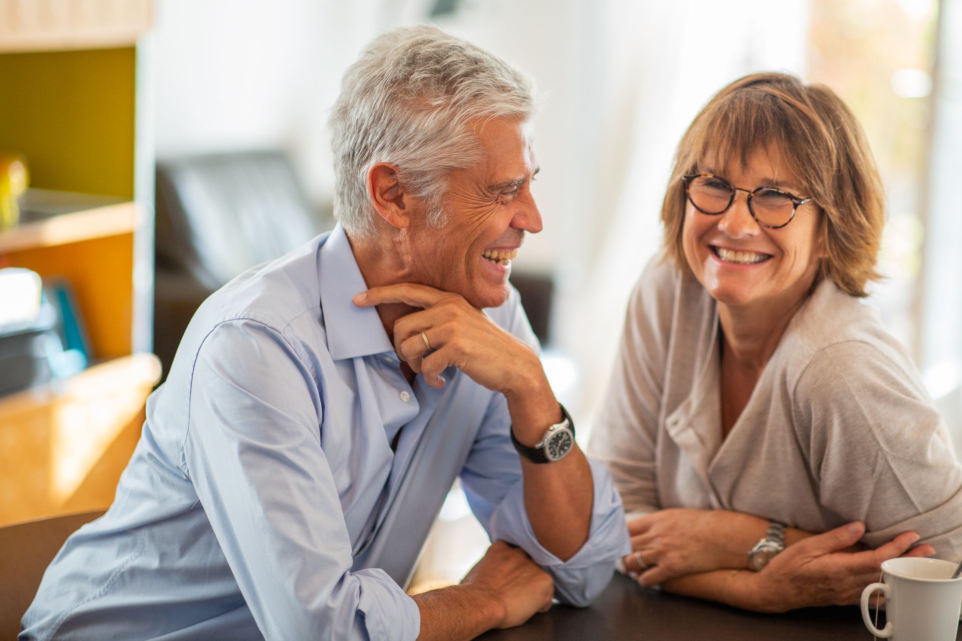 Smiling older couple at a table. Man touches his chin, woman wears glasses, both are indoors.