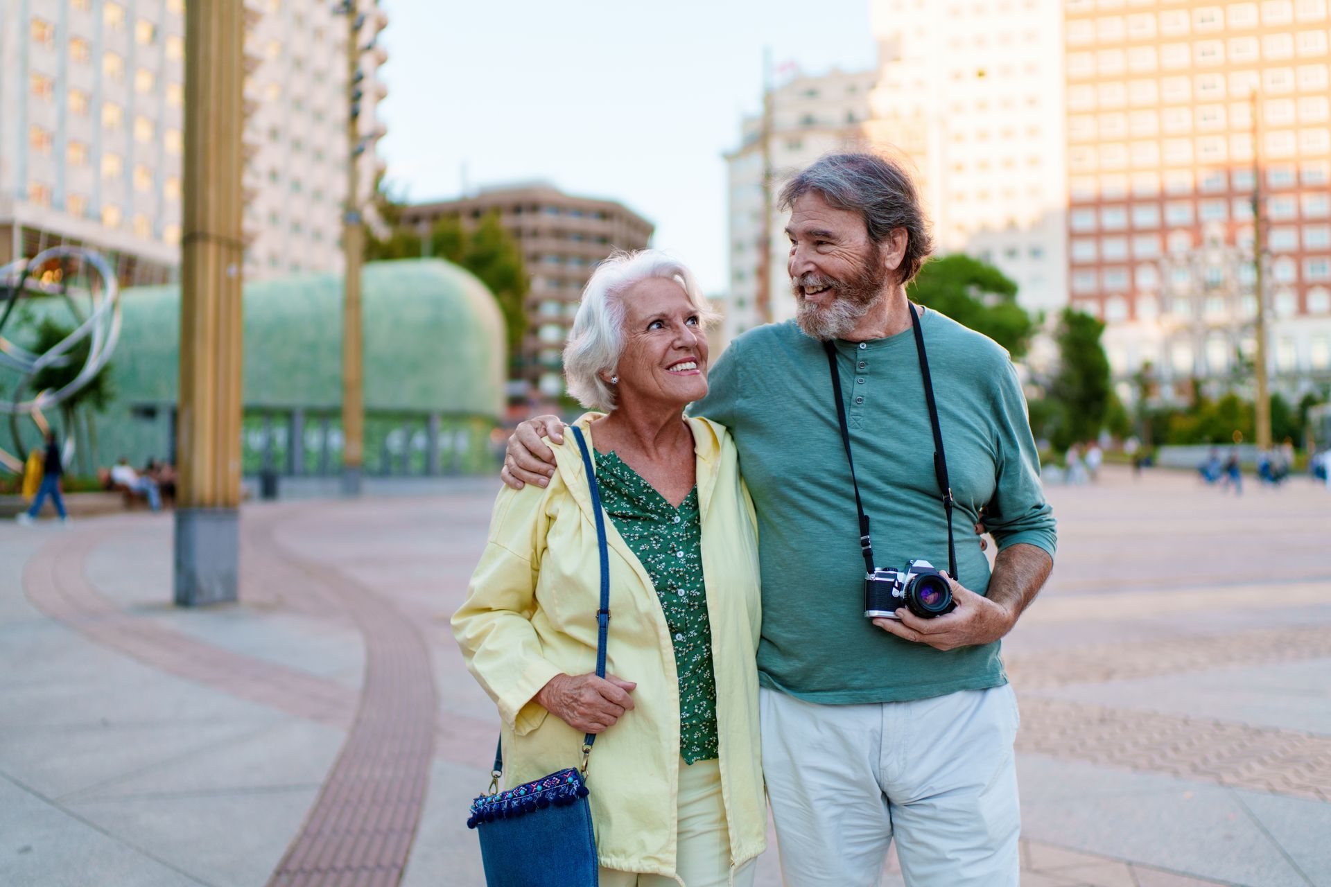 Elderly couple arm-in-arm walking in a city square, smiling. Man holds a camera, woman wears yellow jacket.