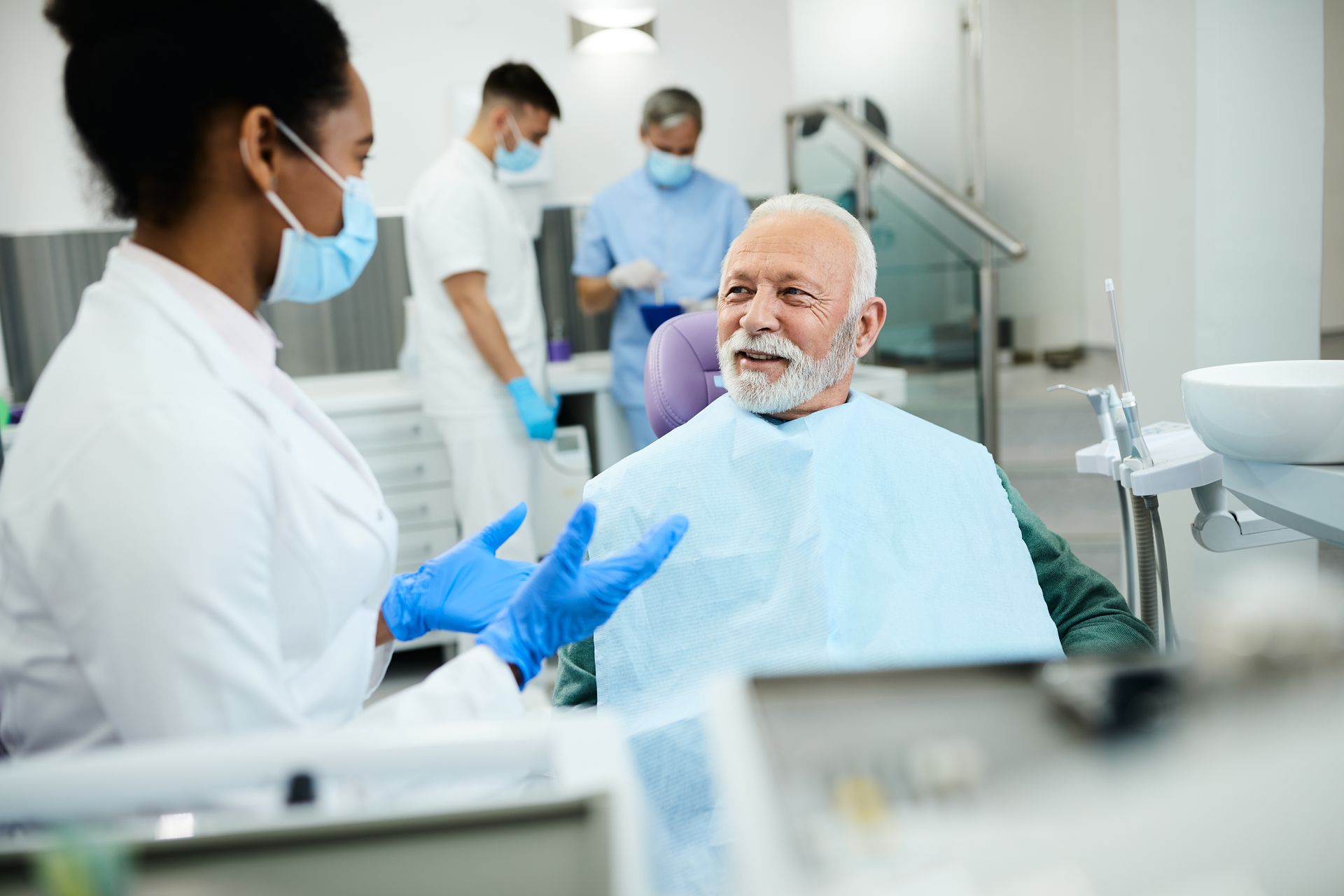 Dentist and patient in a dental office. Dentist wearing gloves and mask gestures. Patient smiles, wearing a bib.