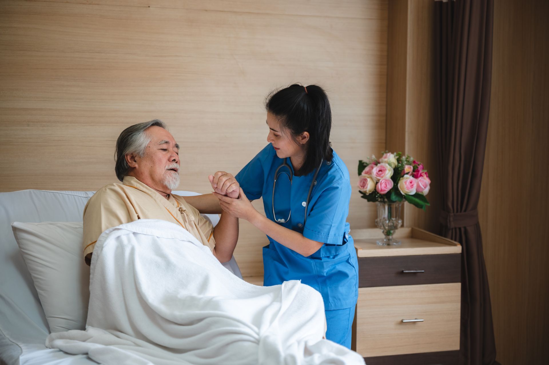 Nurse in blue scrubs examines an elderly man's arm in a hospital room, near a bedside table with flowers.