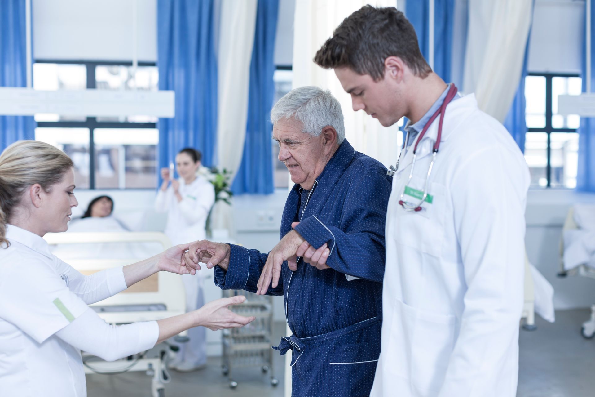Medical staff assist an elderly man walking in a hospital room.