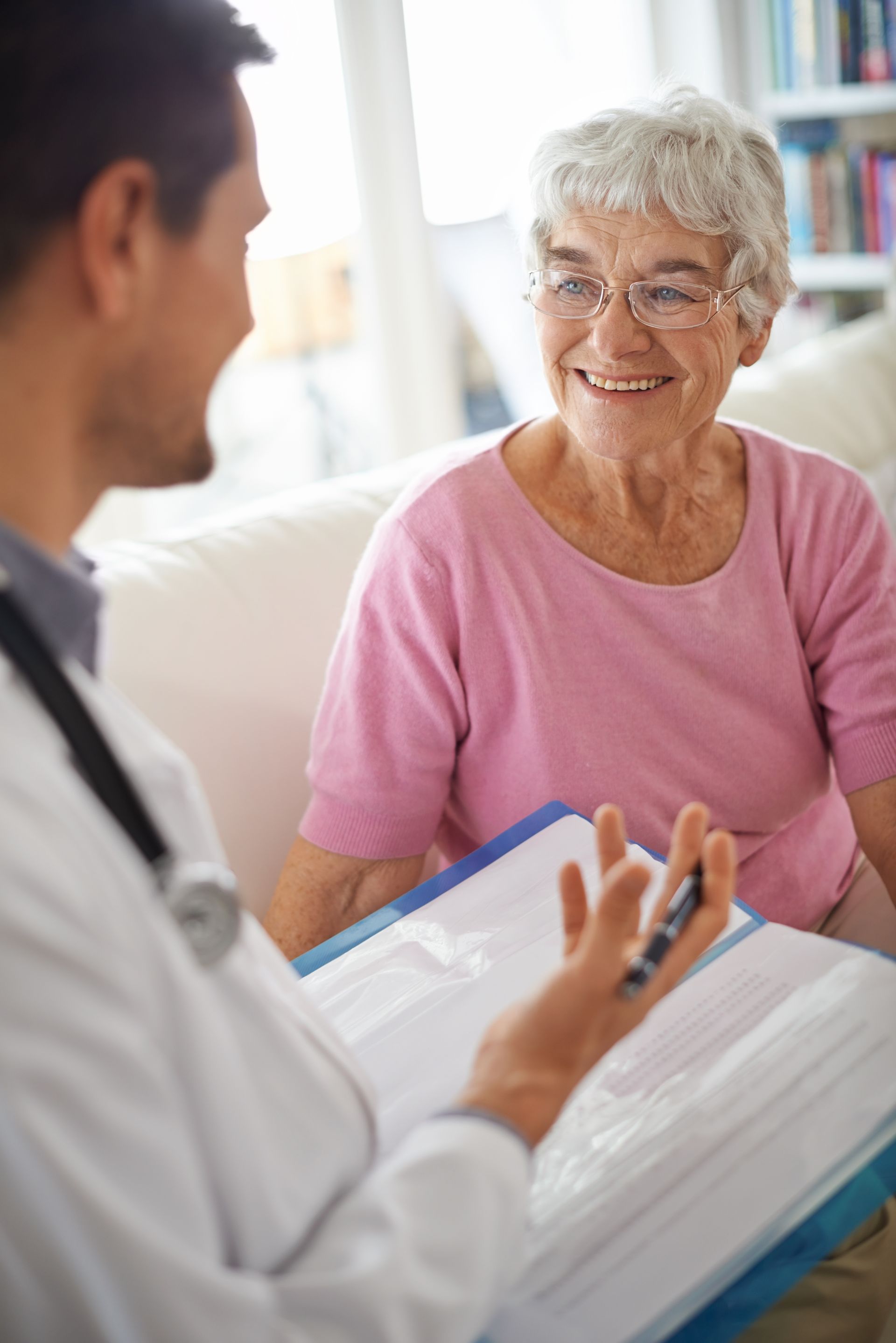 Doctor talking with elderly woman, both smiling. Indoors, woman in pink top, doctor holding paperwork.
