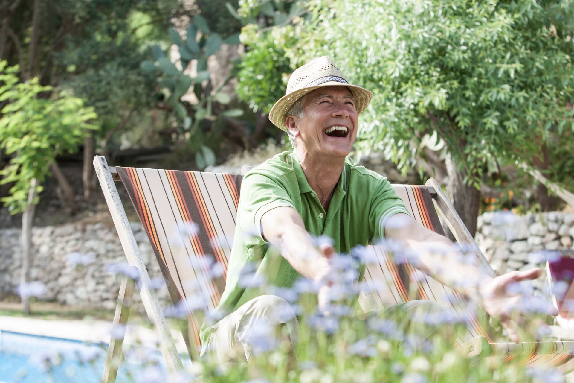 Older man in a straw hat laughs while sitting in a striped chair in a sunny garden.