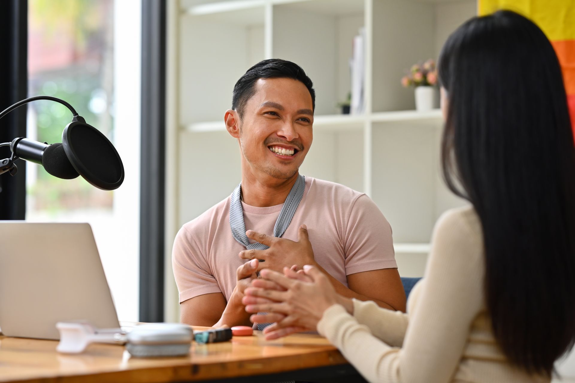 Man and woman in a studio, smiling, gesturing with hands. Microphone and laptop on table, rainbow flag in background.