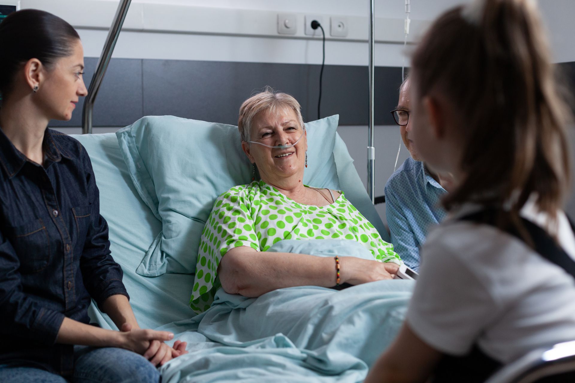 Family visiting a smiling elderly woman in a hospital bed.