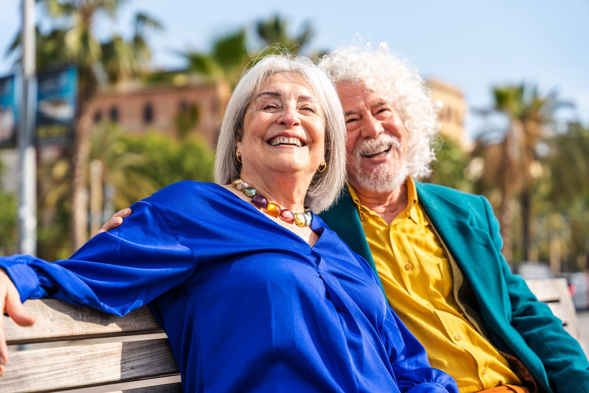 Smiling elderly couple sitting on a bench, woman in blue, man in yellow and teal, palm trees.