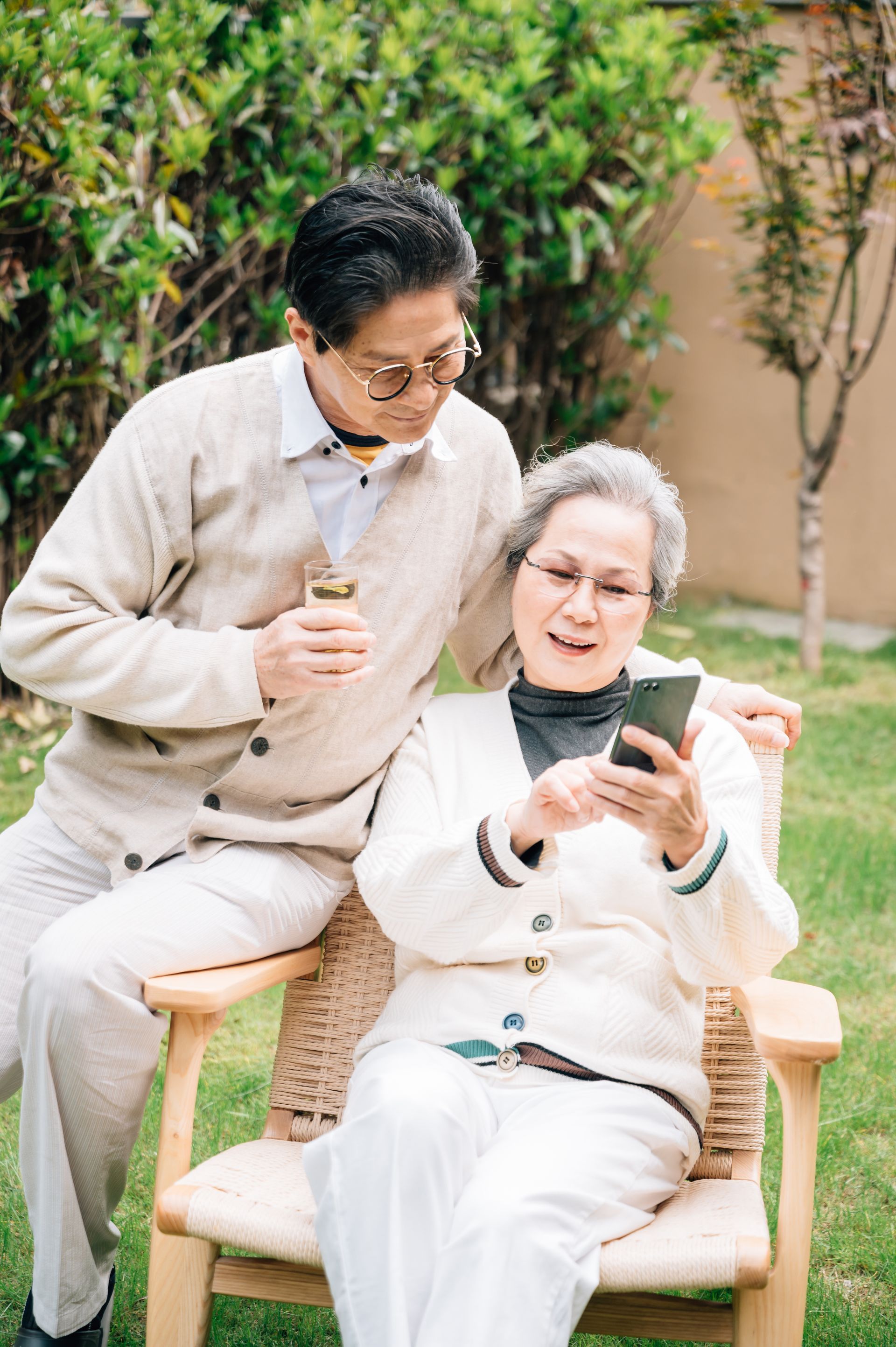 Elderly couple outdoors; woman seated using phone, man looking on, holding drink. Green grass, beige sweaters.