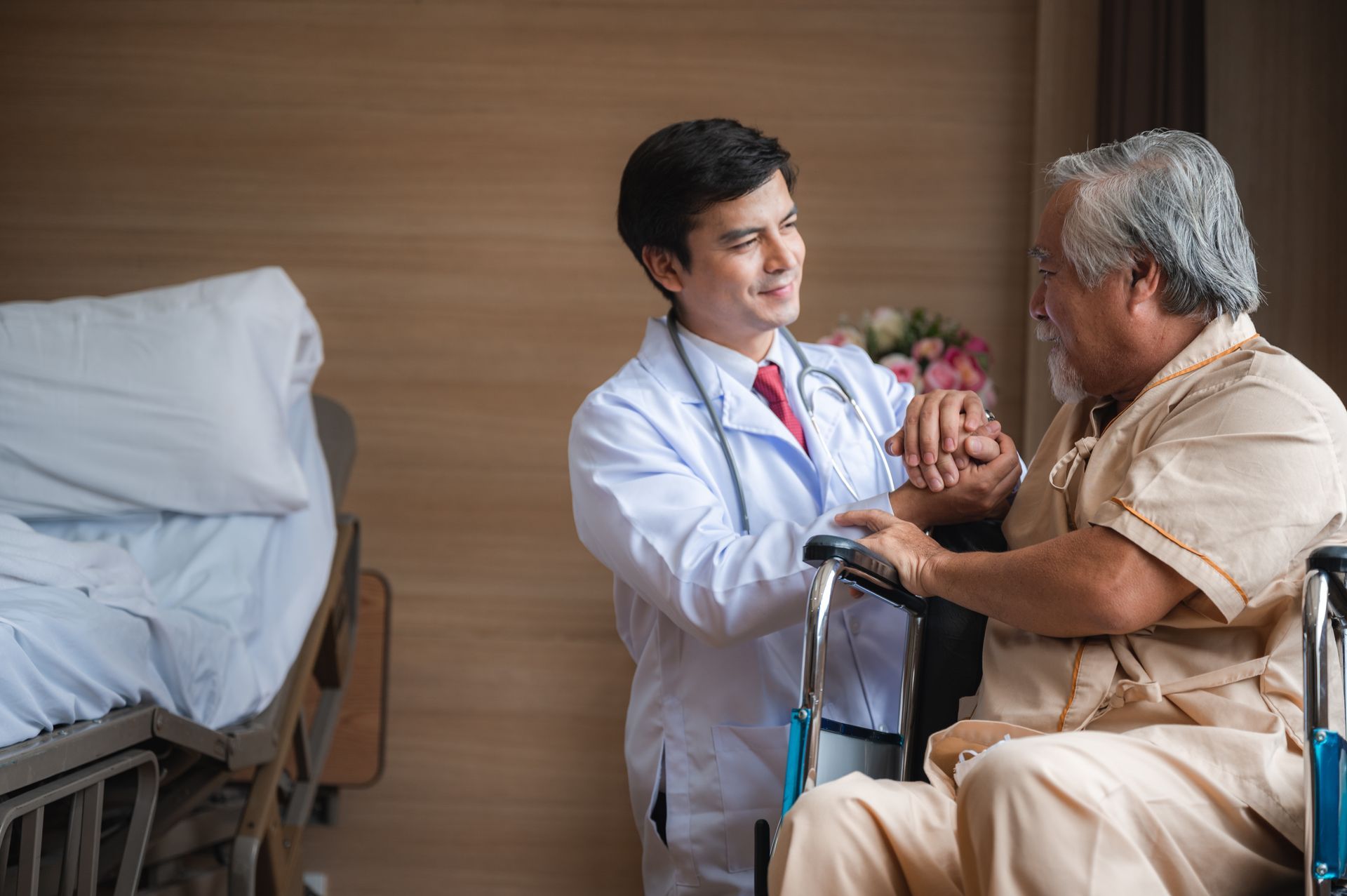 Doctor in white coat helping elderly man in wheelchair, hospital room.